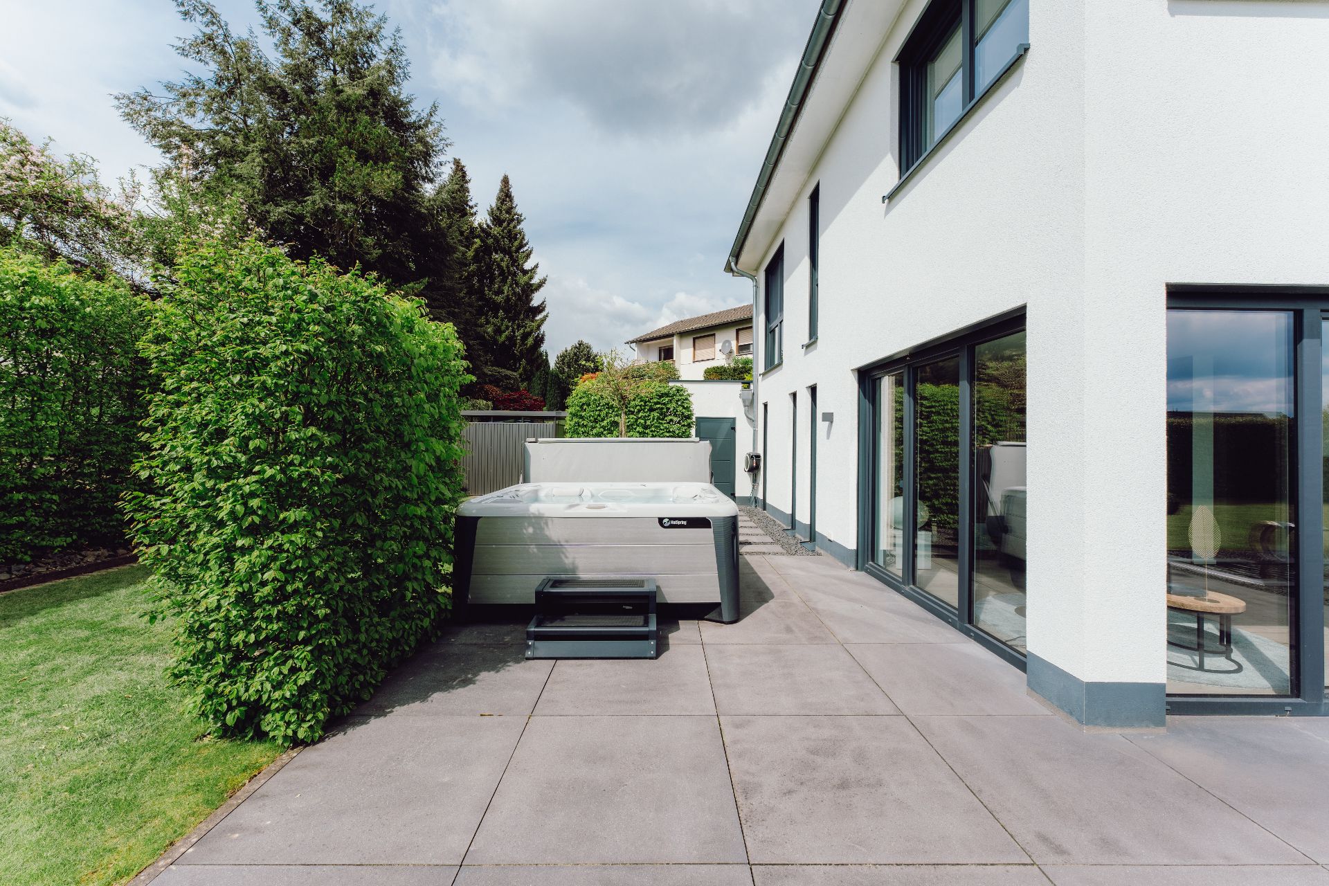 Moderne Terrasse mit Whirlpool neben einem weißen Wohnhaus, umgeben von Hecken.