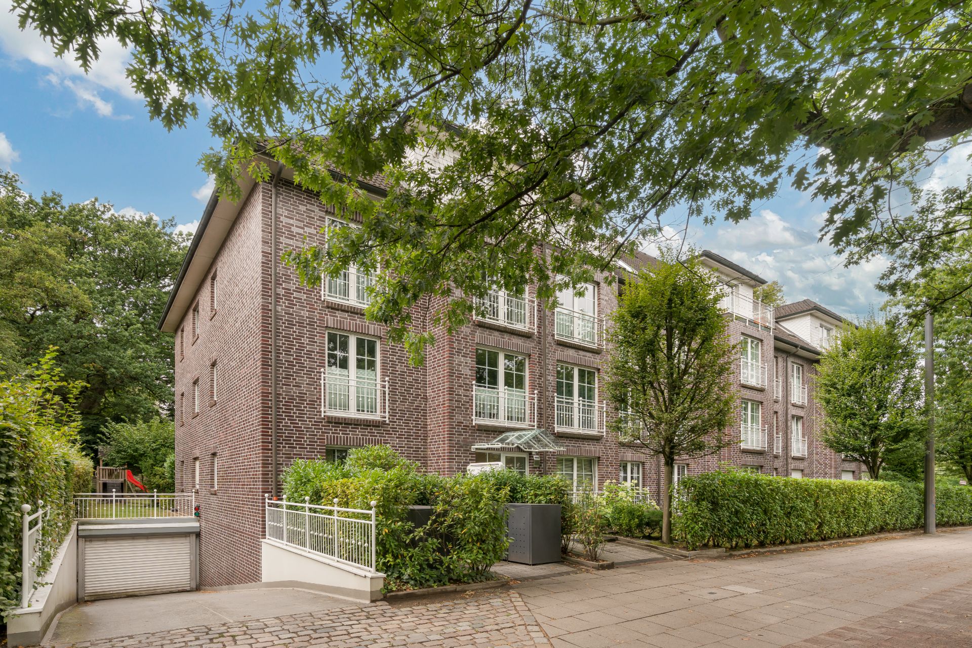 Mehrfamilienhaus aus rotem Backstein mit großen Fenstern, umgeben von Bäumen und Gehwegen.