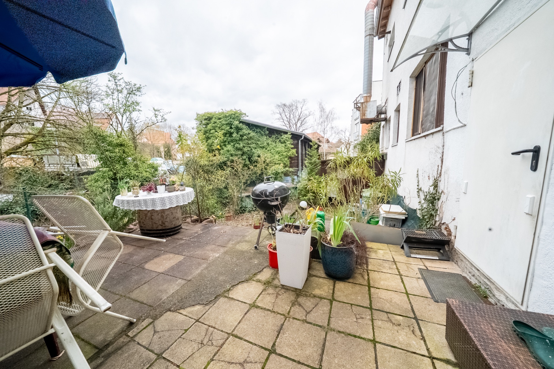 Terrasse mit Gartenmöbeln, Grill, Pflanzkübeln und Bäumen im Hintergrund.
