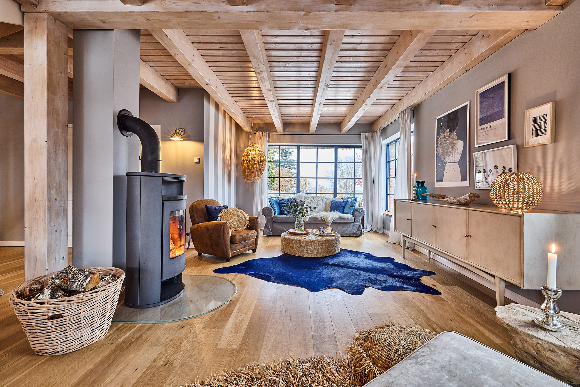 Helles Wohnzimmer mit Holzdecke, Kaminofen, Sofa, Sessel, Teppich und großen Fenstern.