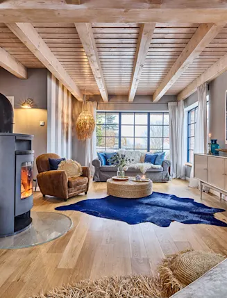 Helles Wohnzimmer mit Holzdecke, Kaminofen, Sofa, Sessel, Teppich und großen Fenstern.