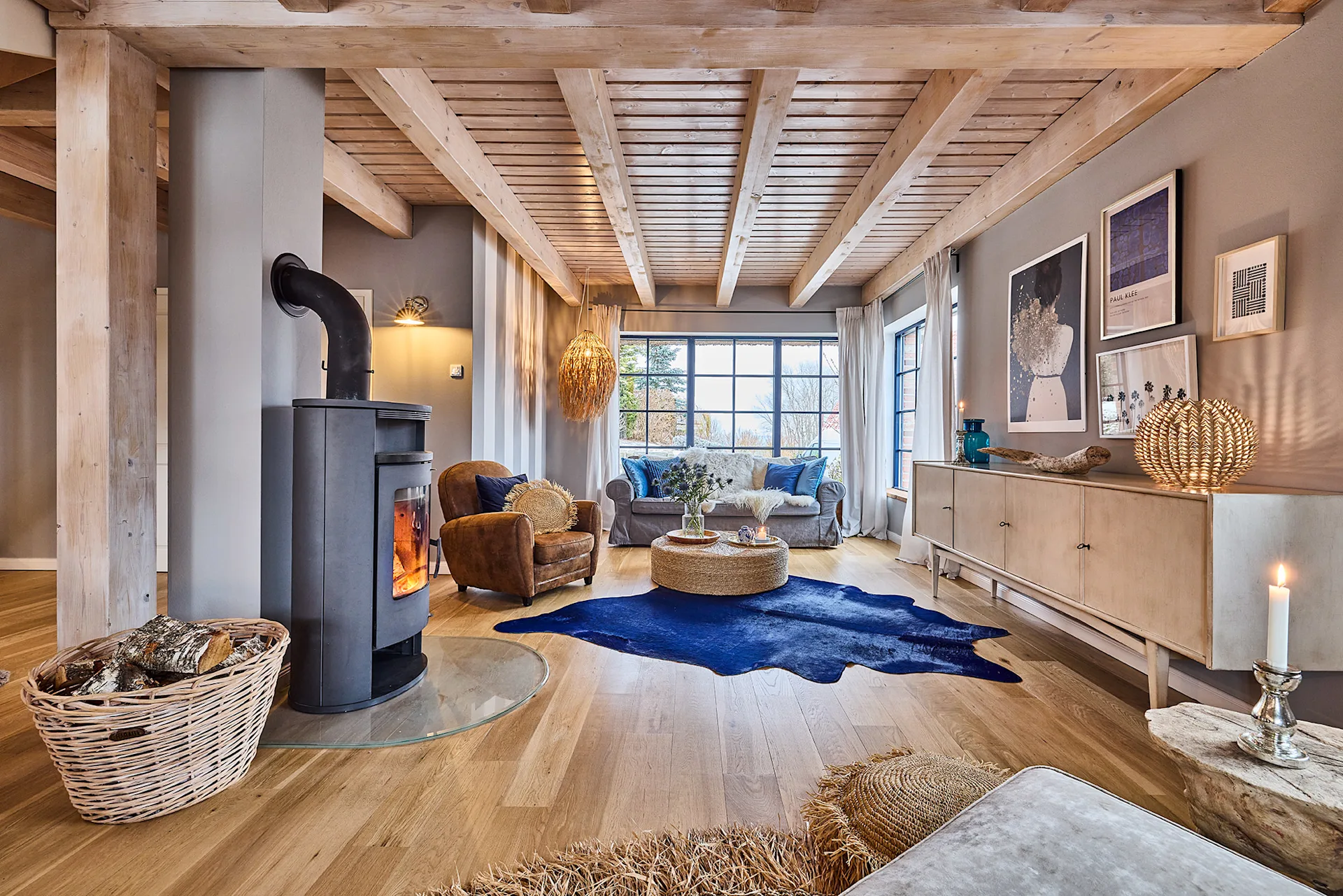 Helles Wohnzimmer mit Holzdecke, Kaminofen, Sofa, Sessel, Teppich und großen Fenstern.