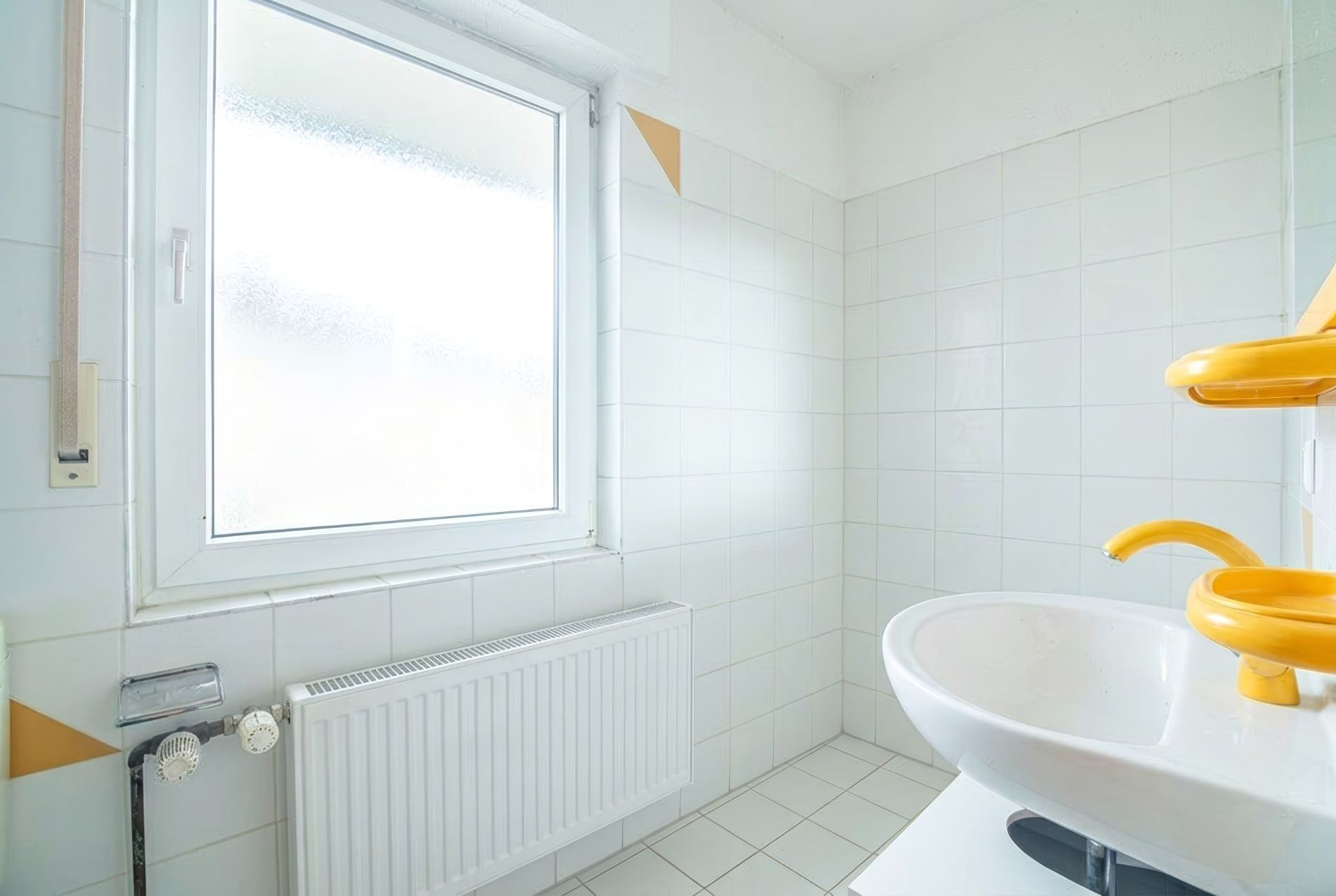 Helles Badezimmer mit weiß gefliesten Wänden, Fenster, Heizkörper und gelbem Waschbecken.