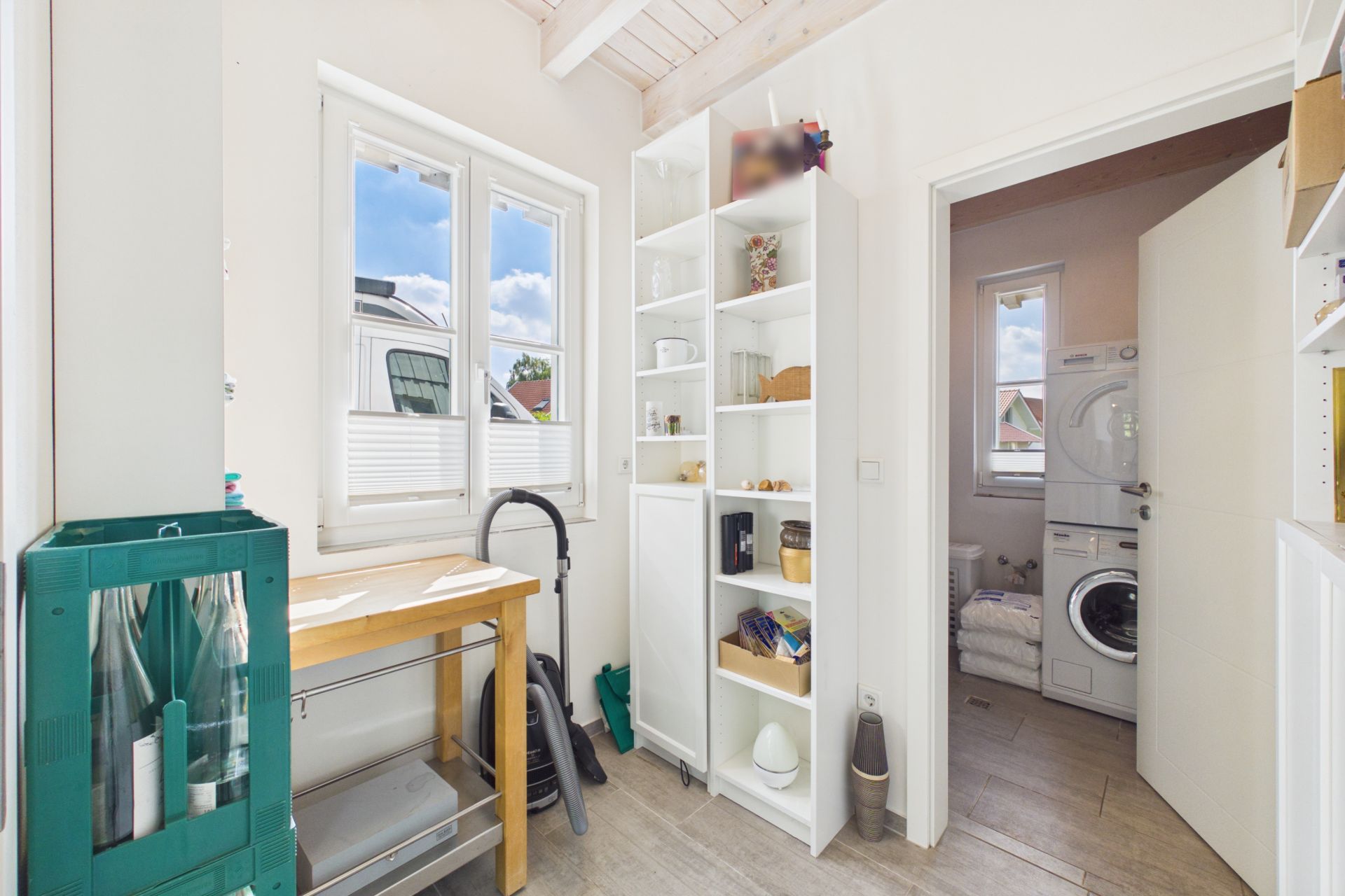 Heller Hauswirtschaftsraum mit Regal, Fenster, grünem Schrank und Zugang zu Raum mit Waschmaschine.
