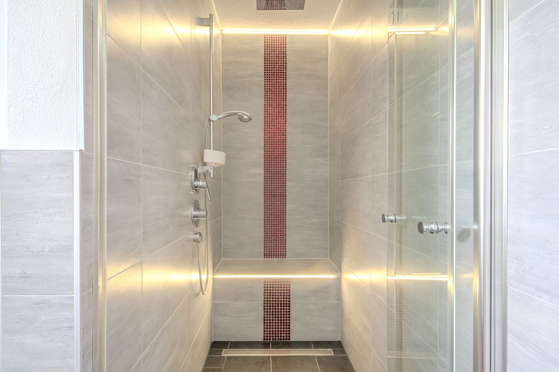 Moderne, beleuchtete Dusche mit grauen Fliesen und Glastür, rote Mosaikleiste an der Rückwand.