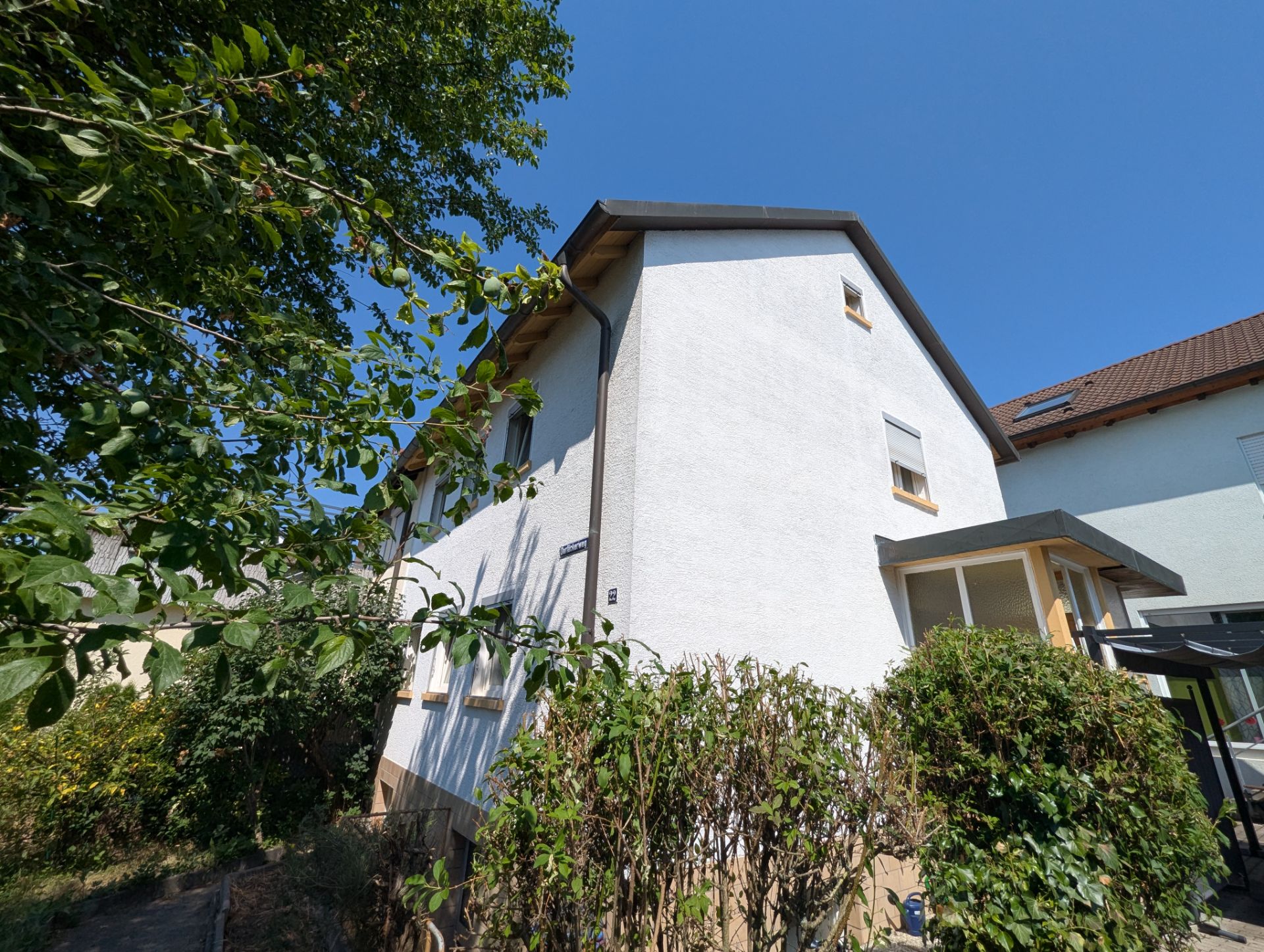 Weißes Wohnhaus mit Satteldach und Garten, umgeben von Büschen und Bäumen an einem sonnigen Tag.
