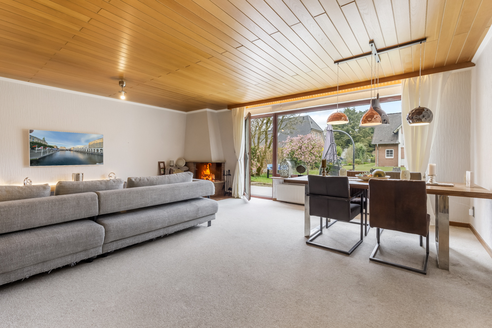 Helles Wohnzimmer mit grauem Sofa, Kamin, Essbereich und großem Fenster mit Blick in den Garten.