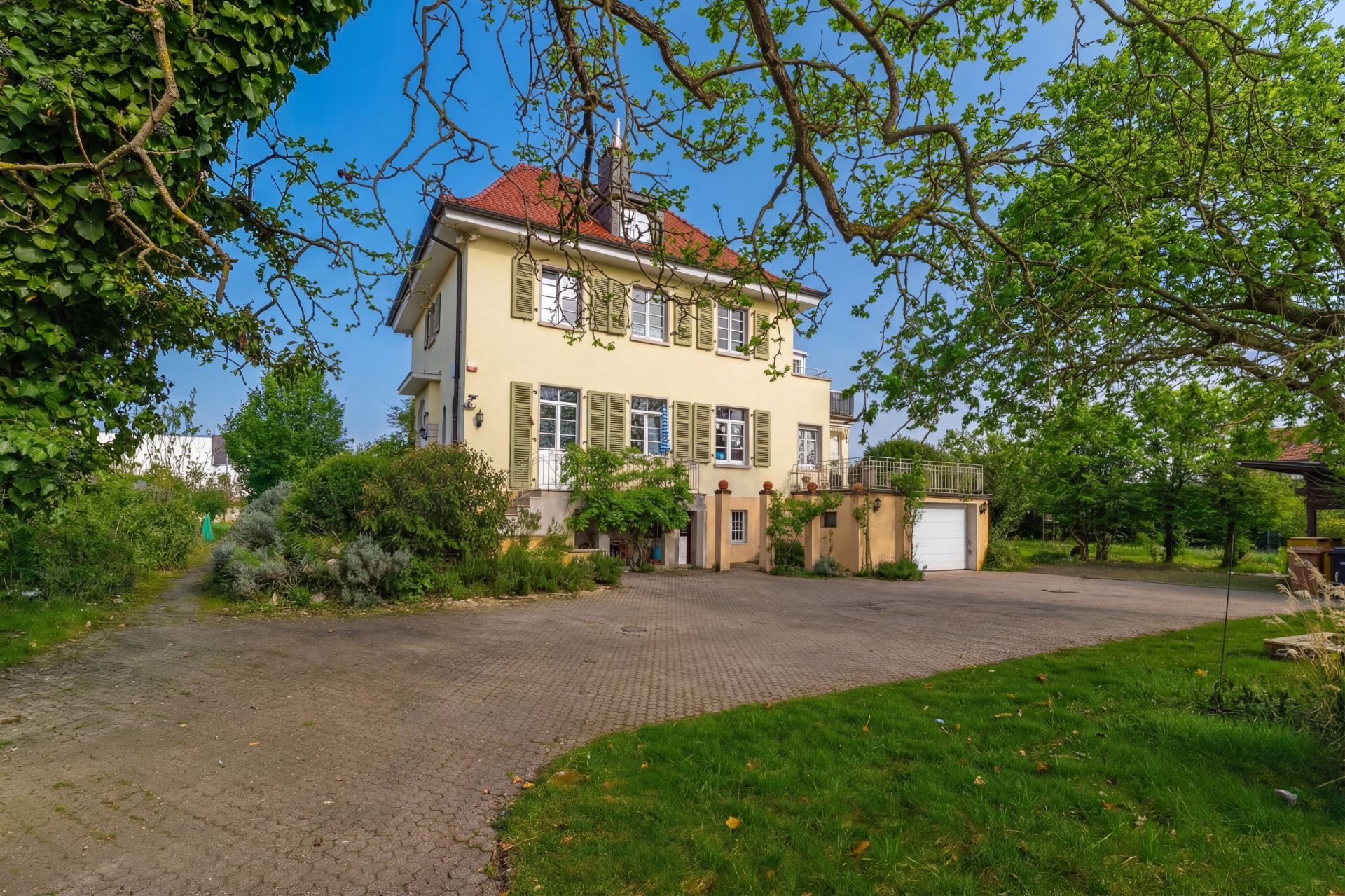 Zweistöckiges Haus mit rotem Dach, grünen Fensterläden und großem Hof, umgeben von Bäumen und Garten.