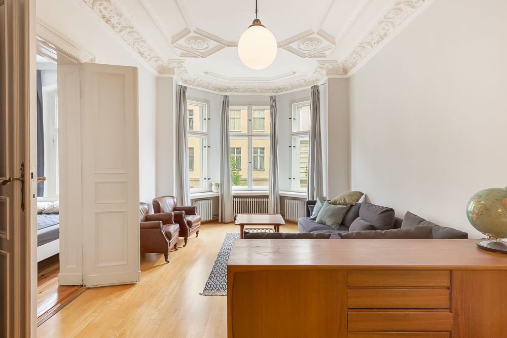 Helles Wohnzimmer mit Stuckdecke, Sofa, Sesseln, Holzboden und bodentiefen Fenstern.