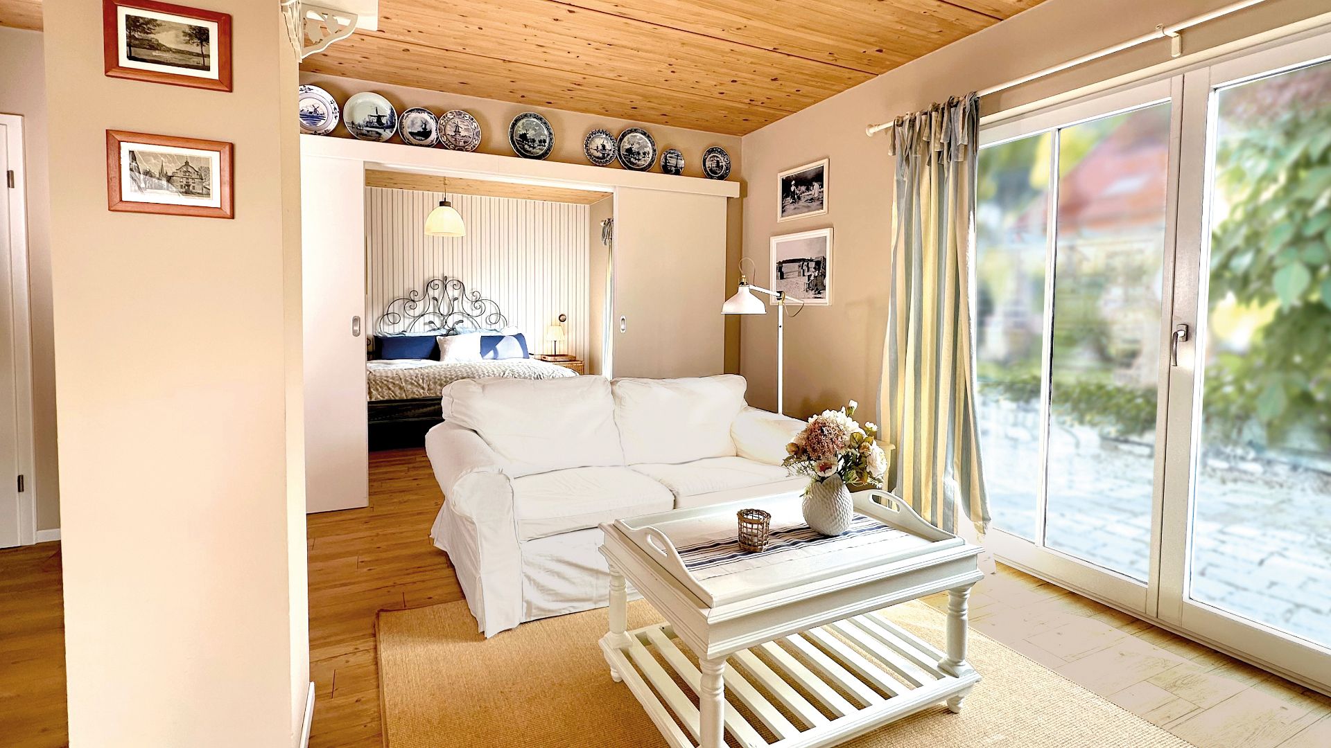 Helles Wohnzimmer mit weißem Sofa, Couchtisch, großen Fenstern und Blick ins Schlafzimmer.