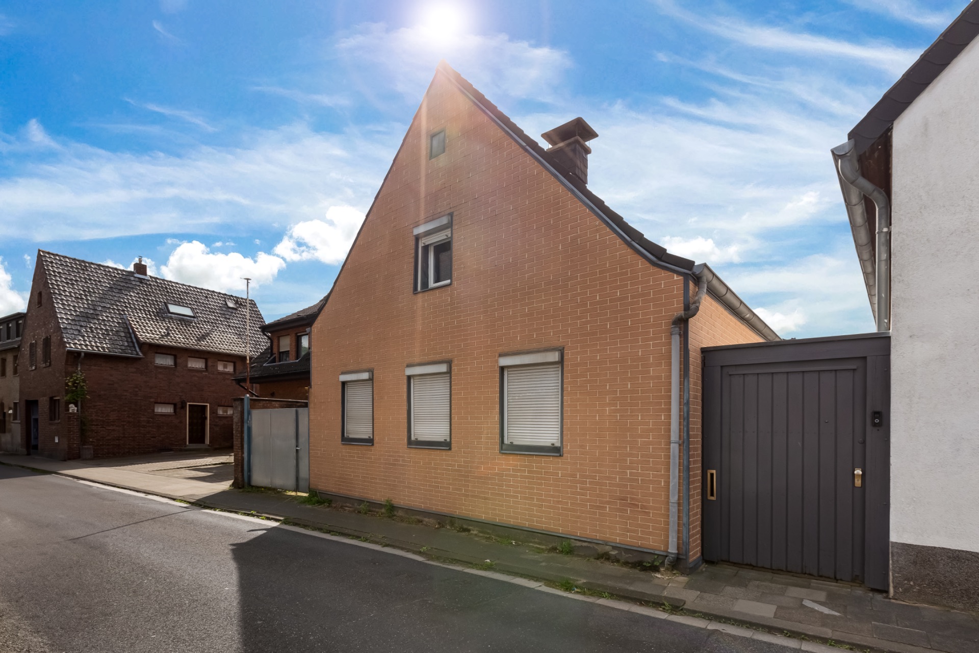 Backsteinwohnhaus mit spitzem Dach, drei Fenstern und angrenzendem Holztor an einer ruhigen Straße.
