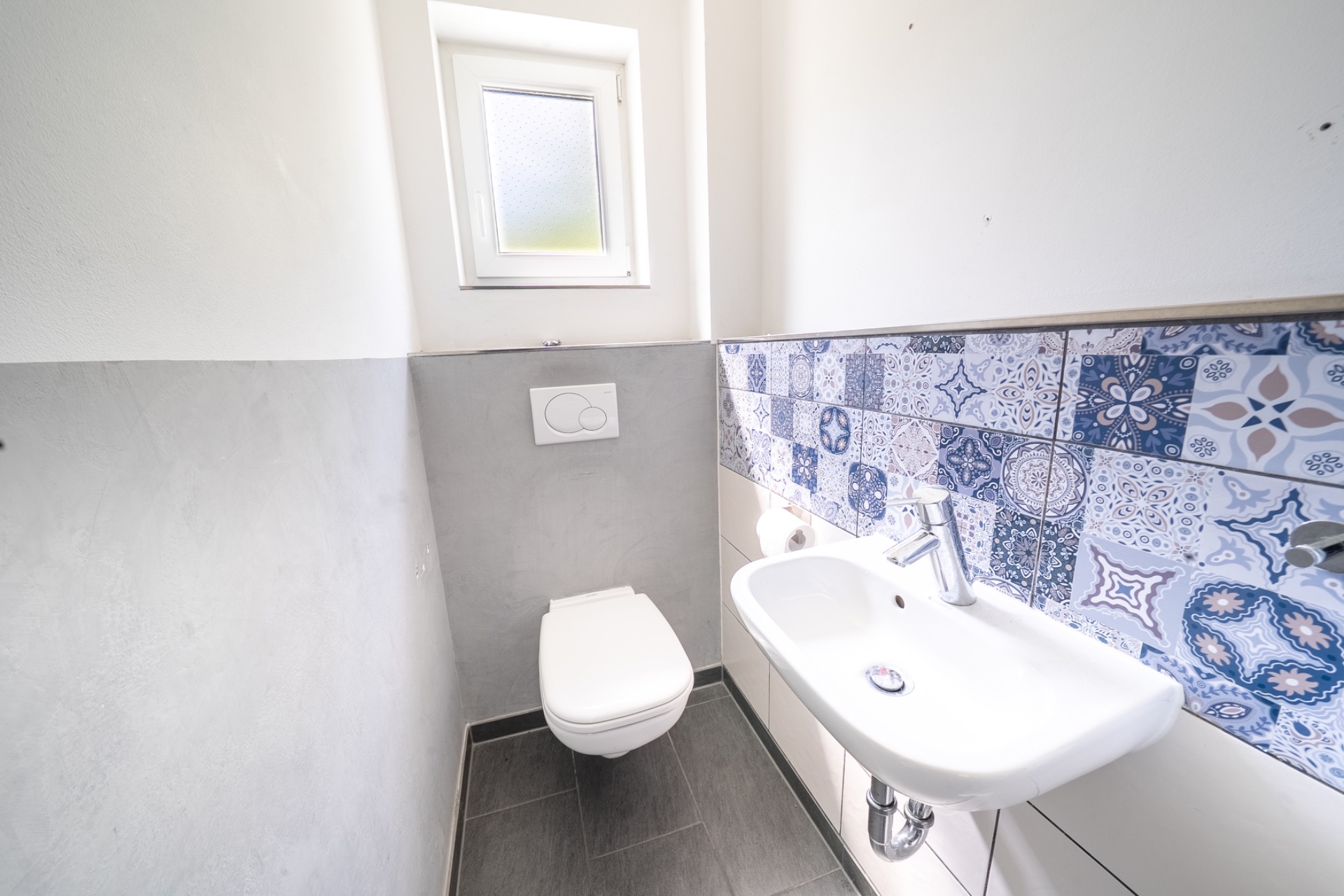 Kleines Bad mit Wand-WC, Waschbecken und gemusterten blauen Fliesen an der Wand.