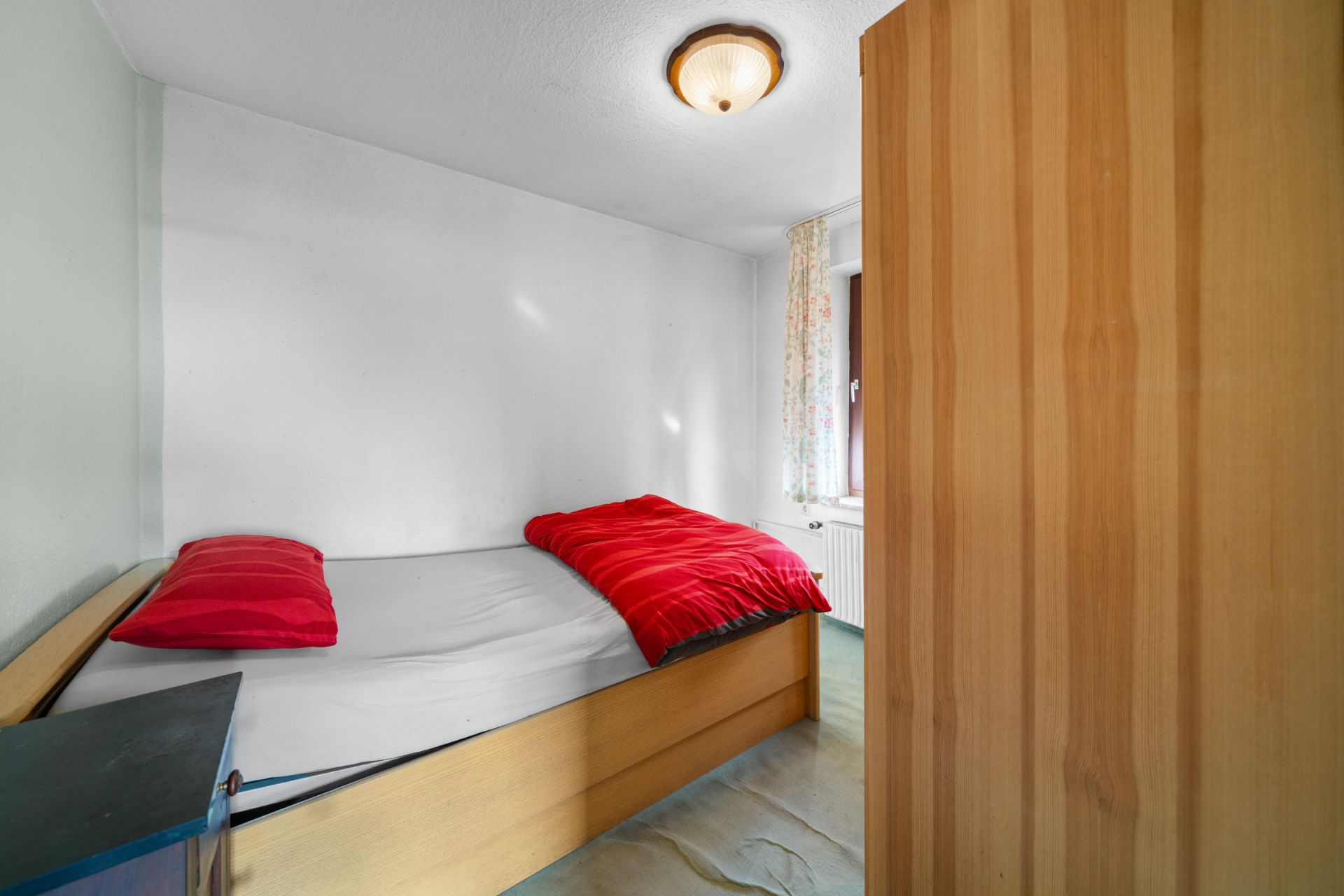 Kleines Schlafzimmer mit Einzelbett, roter Bettdecke, Kissen, Schrank und Fenster mit Vorhang.