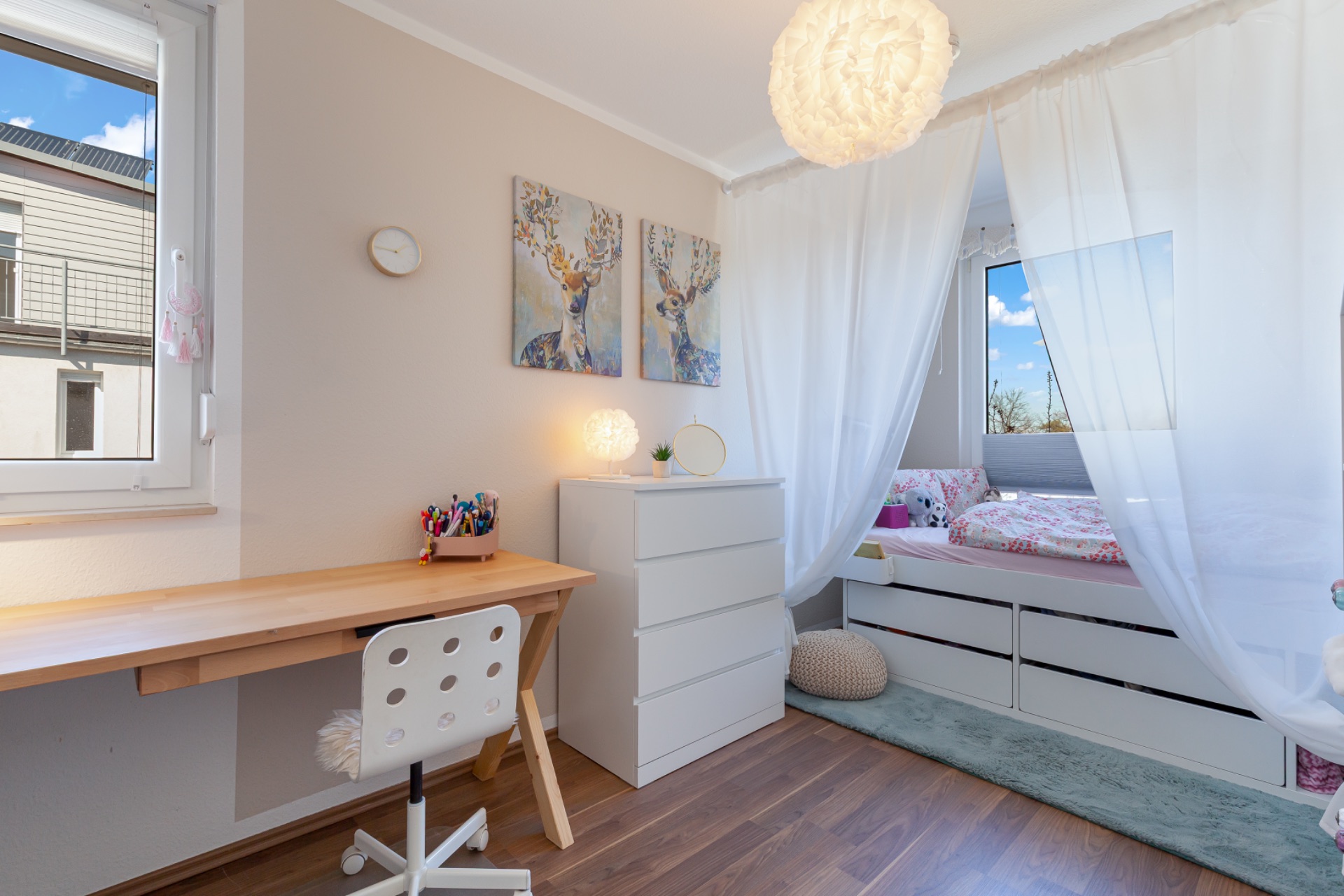 Helles Kinderzimmer mit Schreibtisch, Kommode, Bett und weißen Vorhängen.