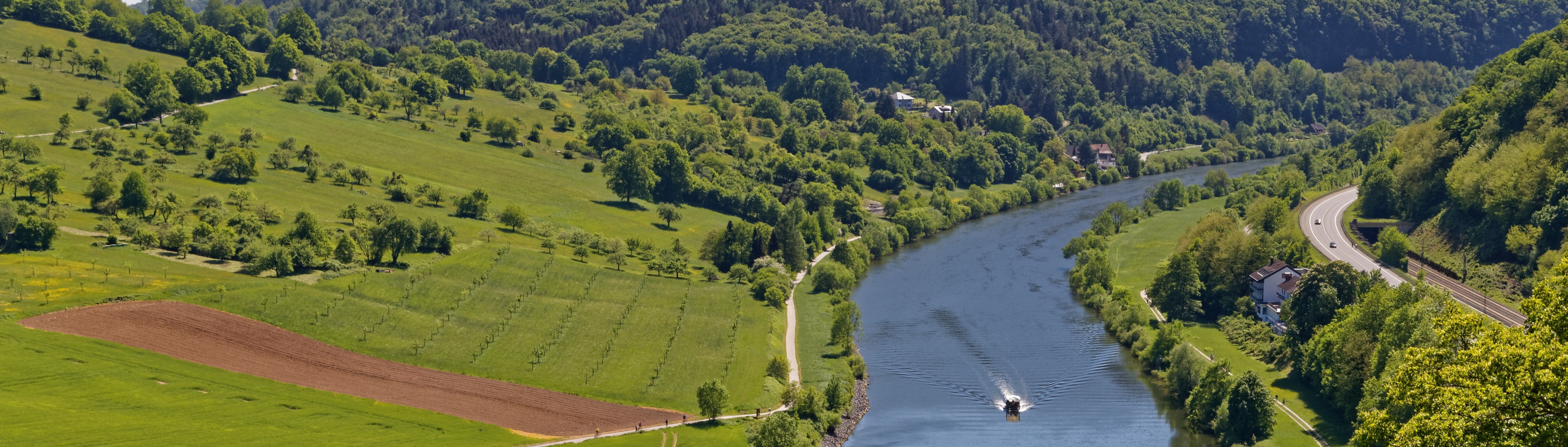 Panorama mit Fluss, grünen Wiesen, Feldern, Bäumen, einem Boot und einer Straße mit Autos.