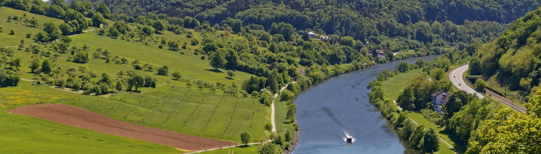 Panorama mit Fluss, grünen Wiesen, Feldern, Bäumen, einem Boot und einer Straße mit Autos.