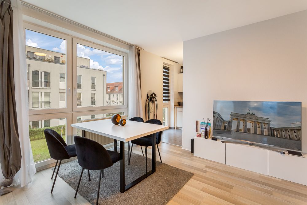 Helles Wohnzimmer mit Esstisch, vier Stühlen, großem Fenster und Fernseher auf weißem Sideboard.