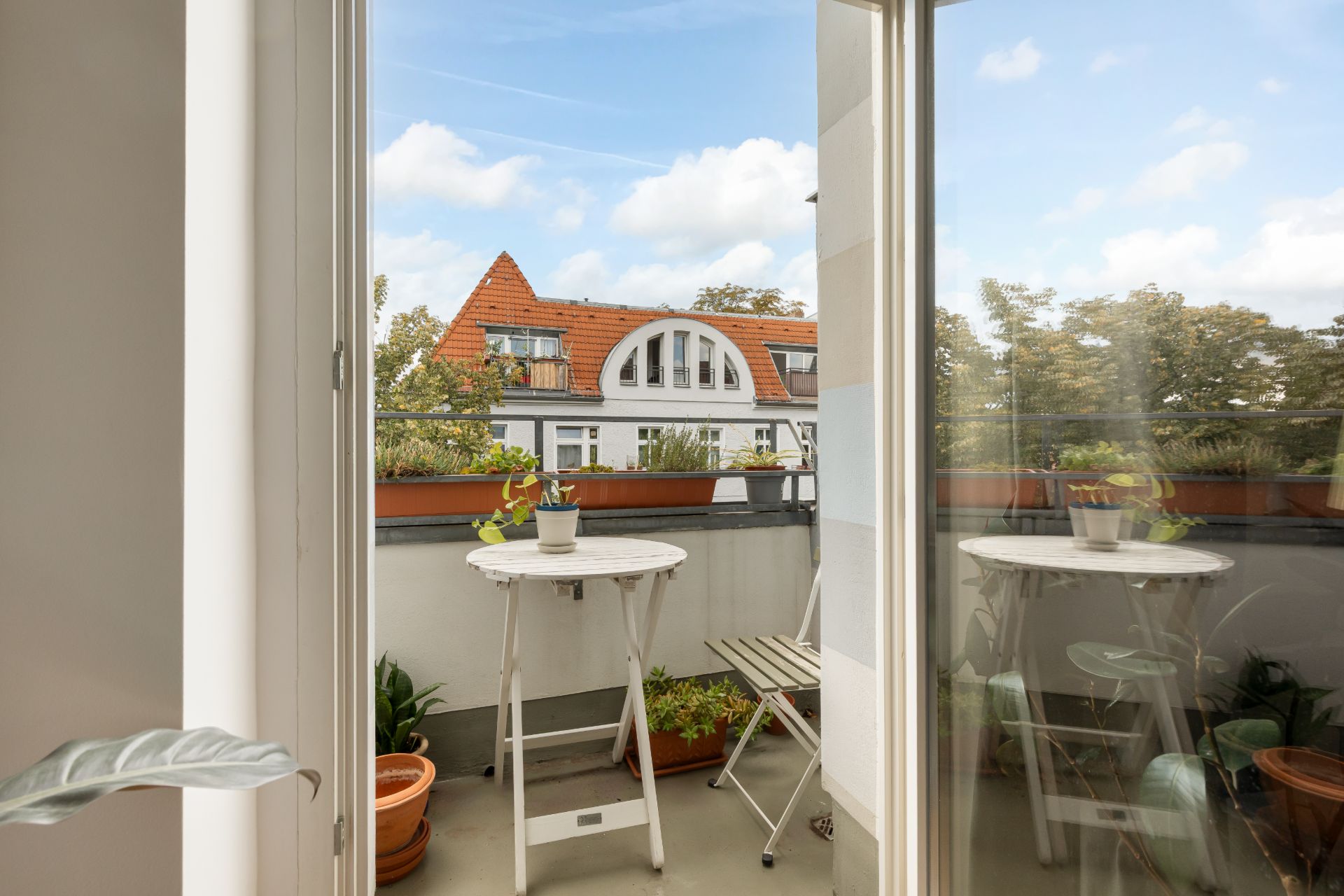 Balkon mit kleinem Tisch, zwei Stühlen und Pflanzen, Blick auf Nachbarhäuser mit roten Dächern.
