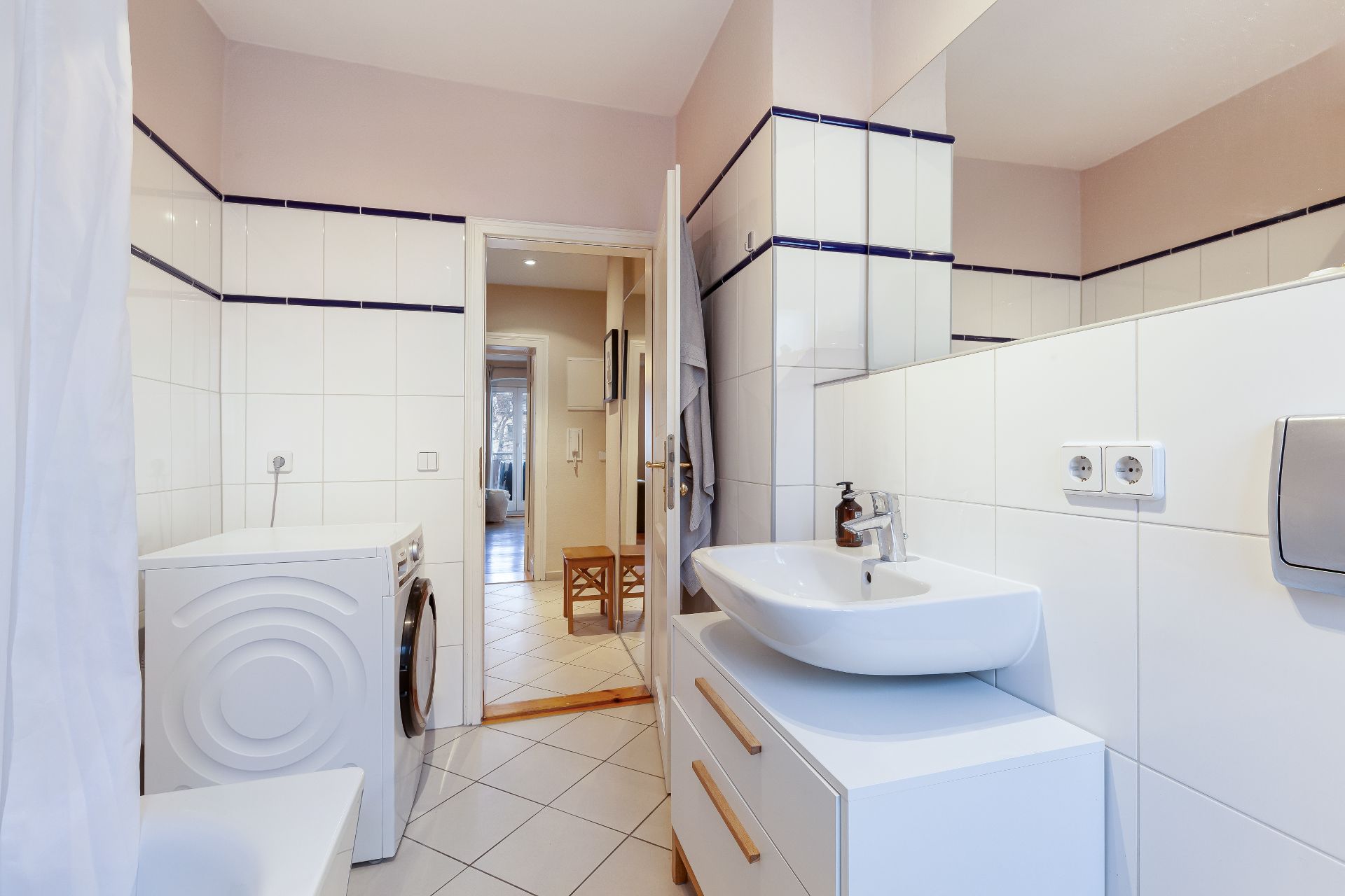 Modernes Badezimmer mit Waschbecken, Waschmaschine, Fliesen und offenstehender Tür zum Flur.