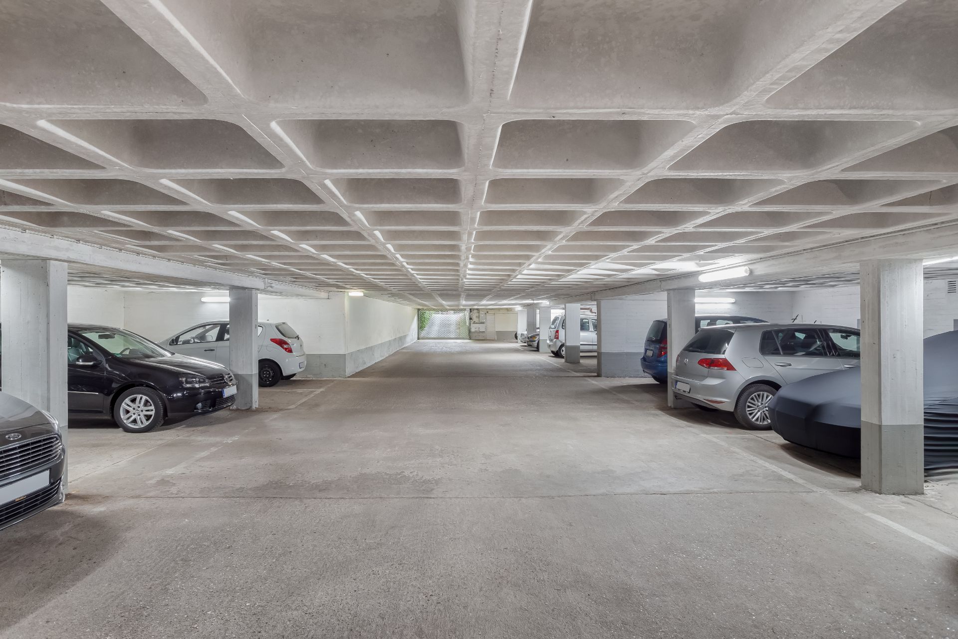 Tiefgaragenstellplätze mit geparkten Autos und Betonsäulen, beleuchtet durch Deckenlampen.