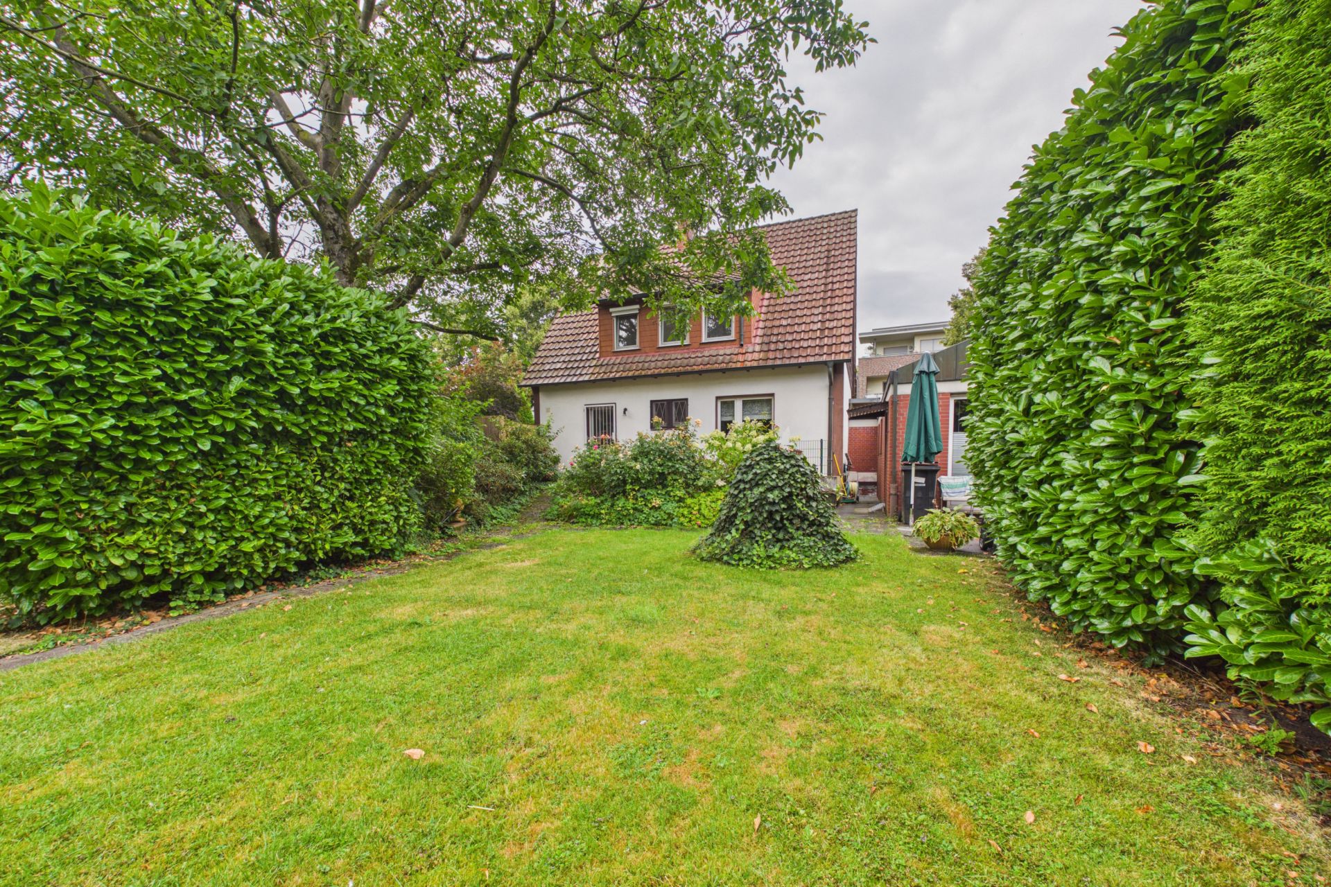 Gepflegter Garten mit Rasen, Sträuchern und kleinem Haus im Hintergrund.