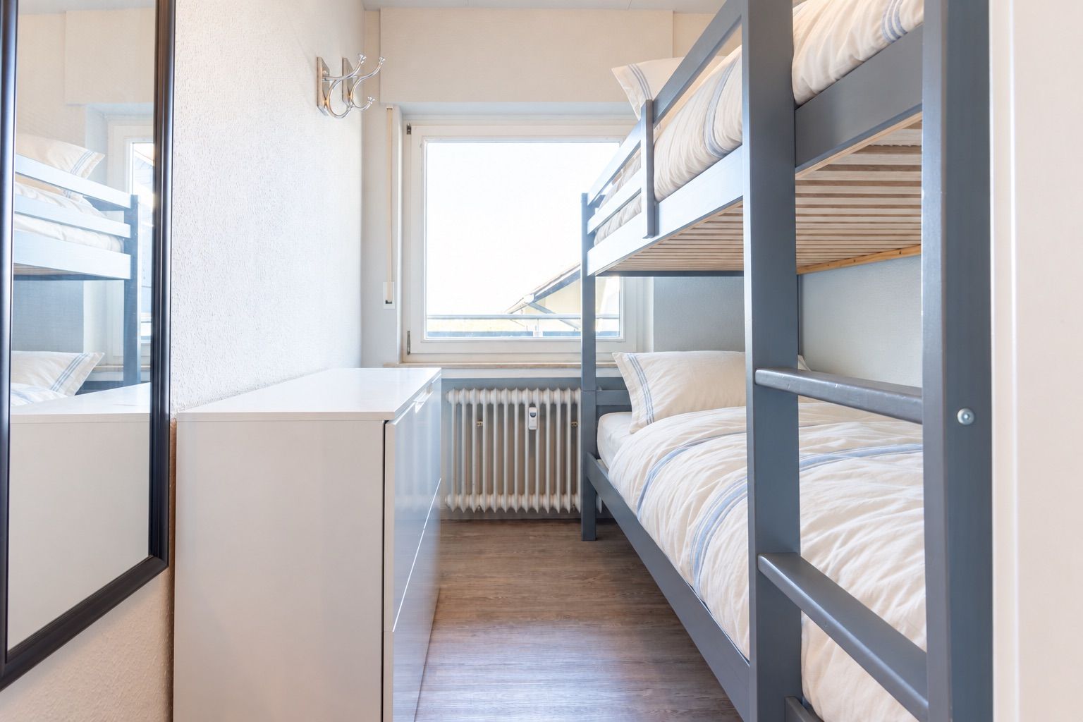 Zimmer mit Etagenbett, Kommode, Spiegel und großem Fenster auf Holzboden.