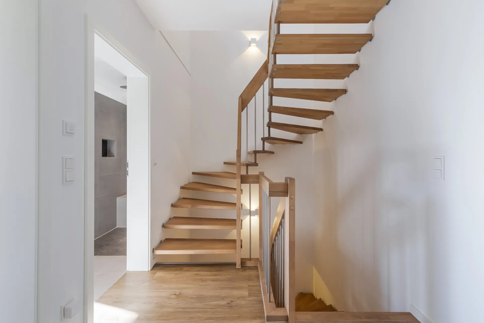 Helle Diele mit moderner Holztreppe und Blick in ein angrenzendes Badezimmer.