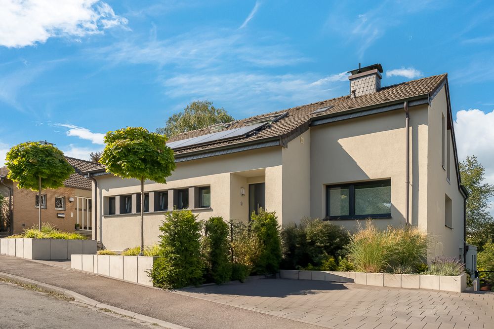 Modernes Einfamilienhaus mit Flachdach, großen Fenstern und Vorgarten mit Bäumen und Sträuchern.