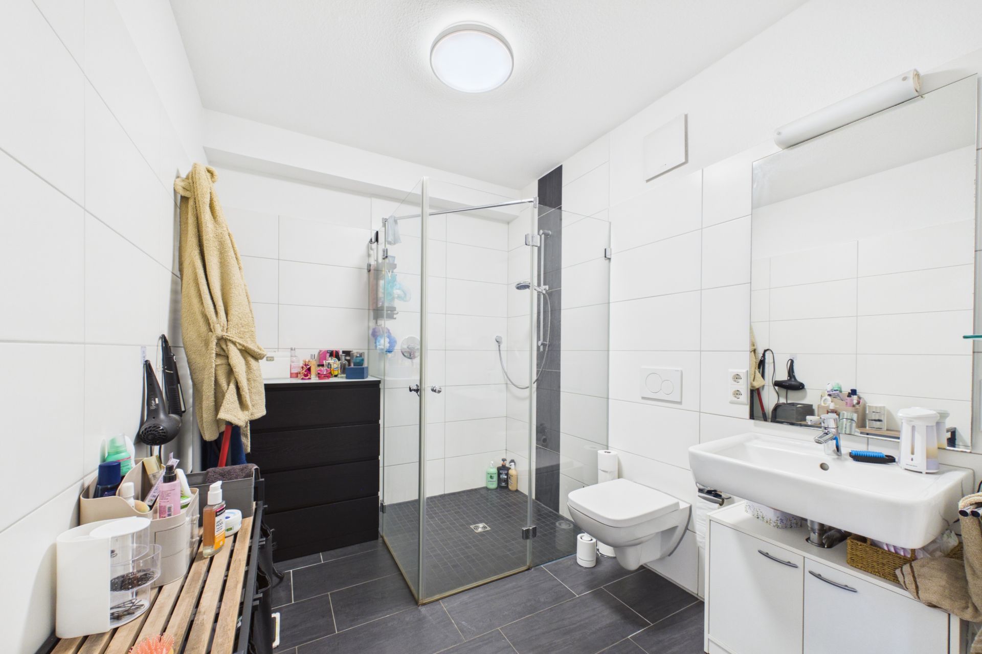Modernes Badezimmer mit Dusche, Waschbecken, WC und Ablagen, weiße Fliesen und dunkler Boden.