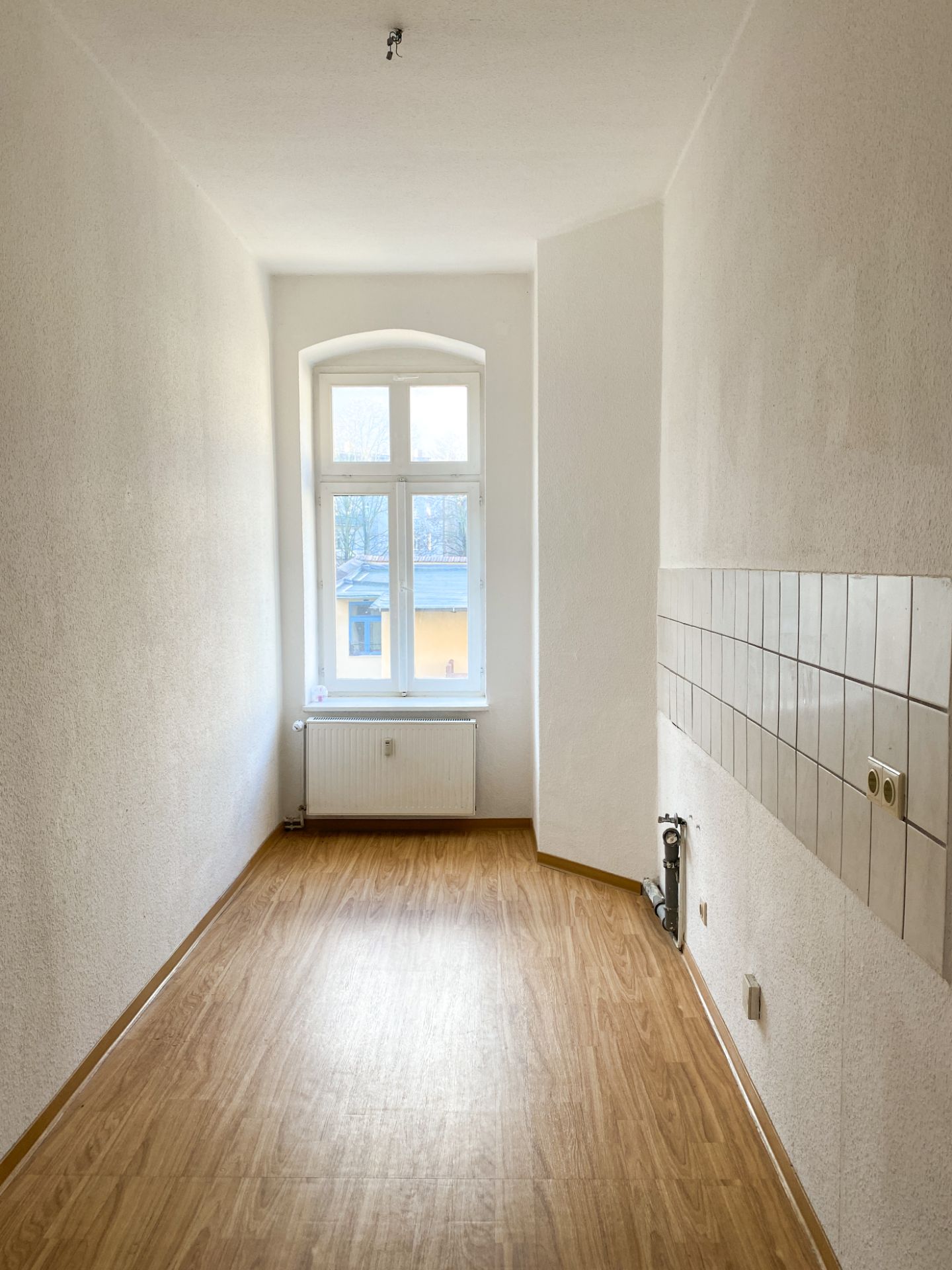 Langer, schmaler Raum mit Fenster, Heizung, Fliesen an der Wand und hellem Boden.