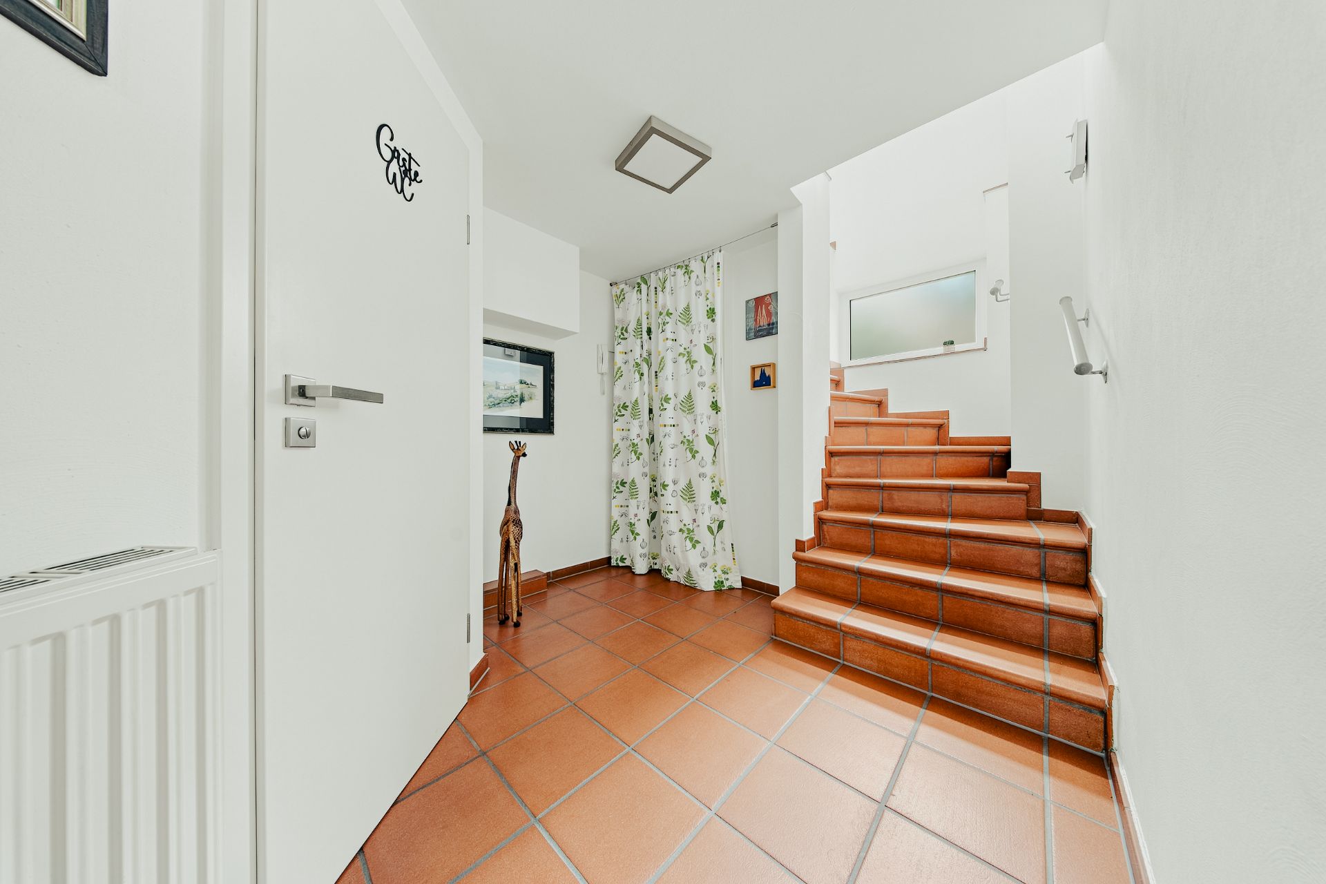 Flur mit braunen Fliesen, Treppe, weißer Tür und Vorhang mit Blattmuster.