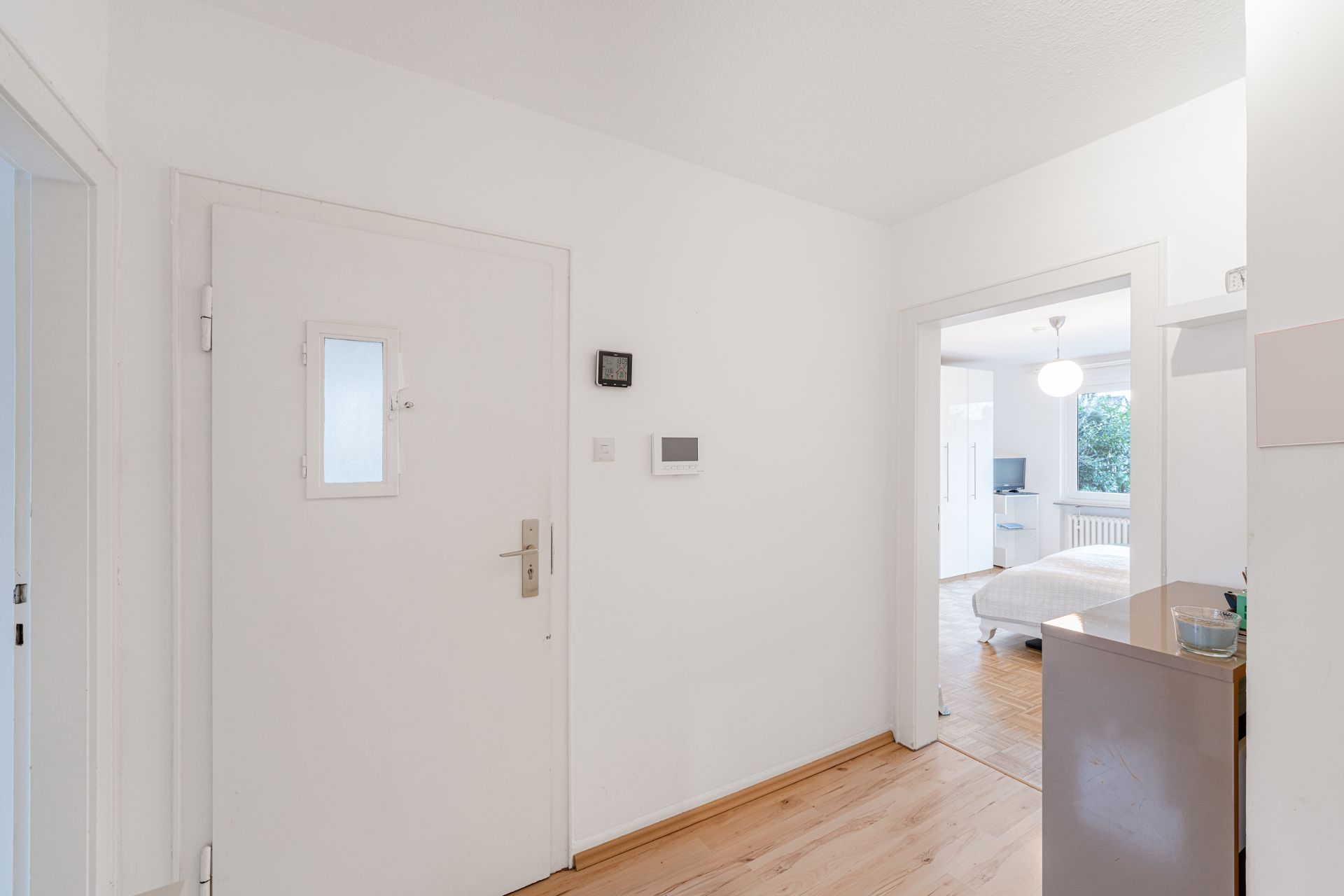 Heller Flur mit weißer Eingangstür, Holzboden und Blick in ein Schlafzimmer.