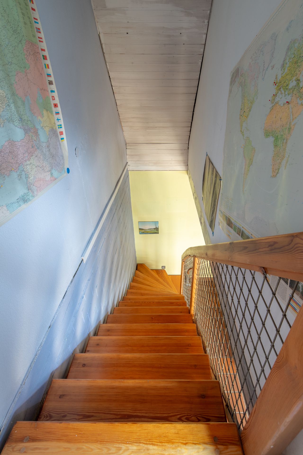 Holztreppe mit Geländer, Karten an den Wänden, Blick nach unten in einen Flur.