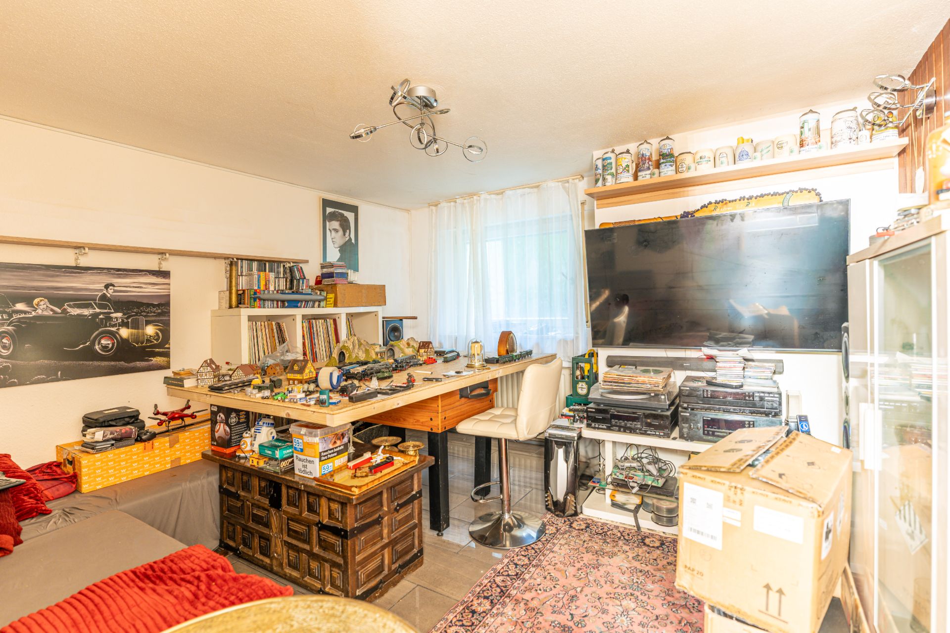 Wohnzimmer mit Sofa, großem Fernseher, Bücherregal und Schreibtisch mit Sammelobjekten.