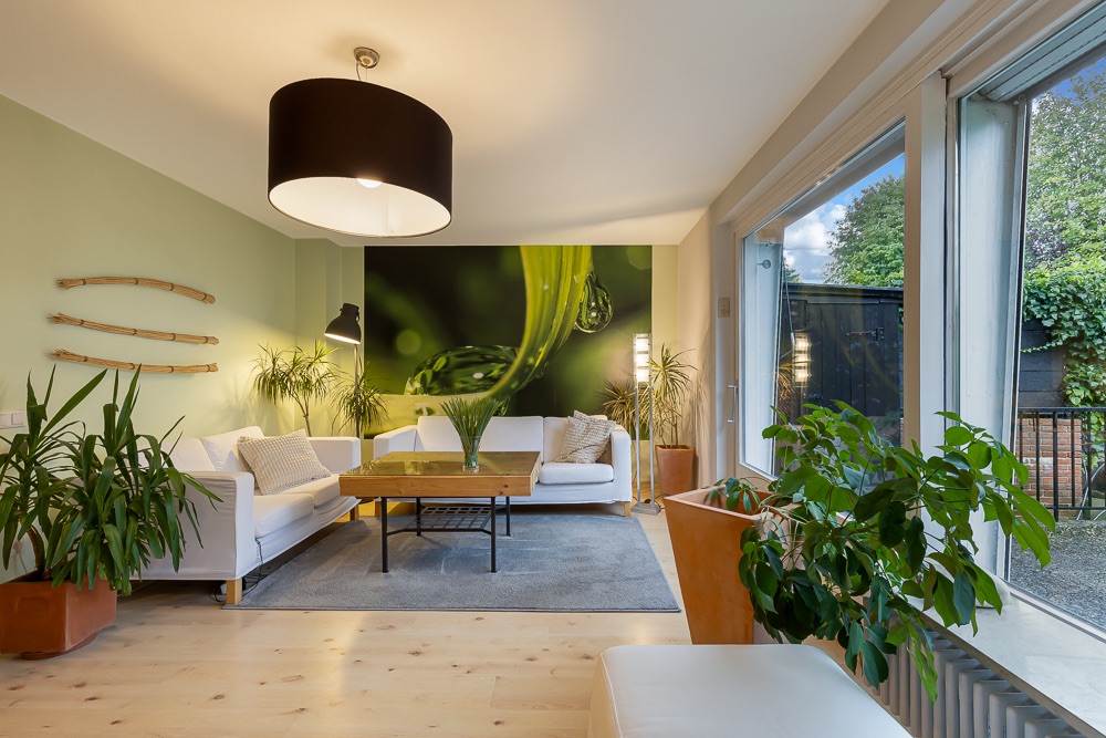 Helles Wohnzimmer mit weißen Sofas, Holztisch, großen Pflanzen und Wandbild mit Naturmotiv.