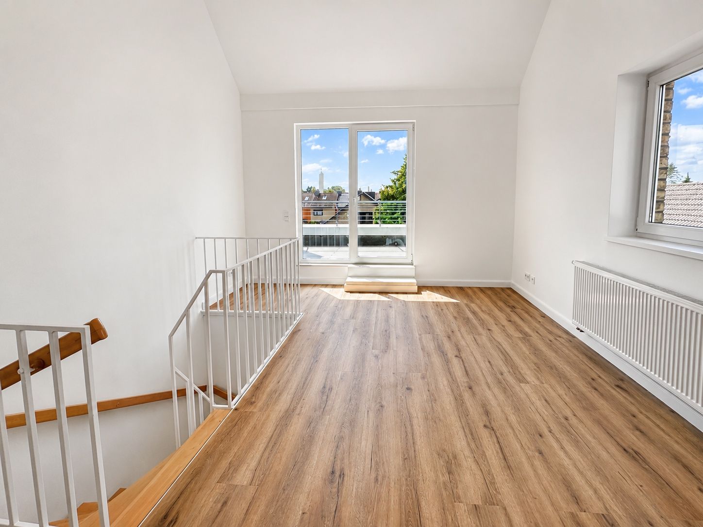 Helles Zimmer mit Holzfußboden, Balkontür, Fenster und Treppe.