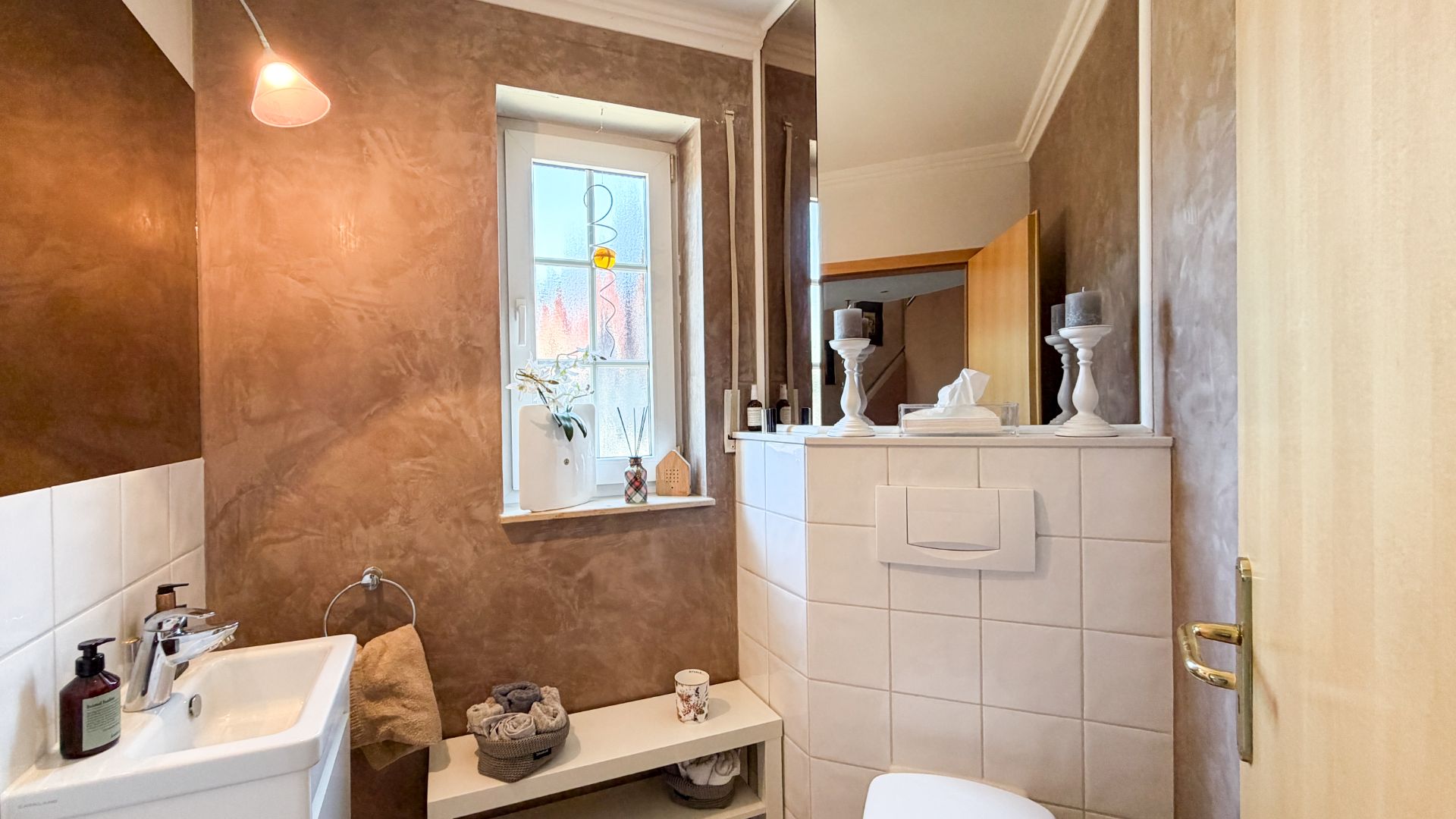 Kleines Badezimmer mit Waschbecken, Toilette, Fenster und dekorativen Elementen auf Ablage.