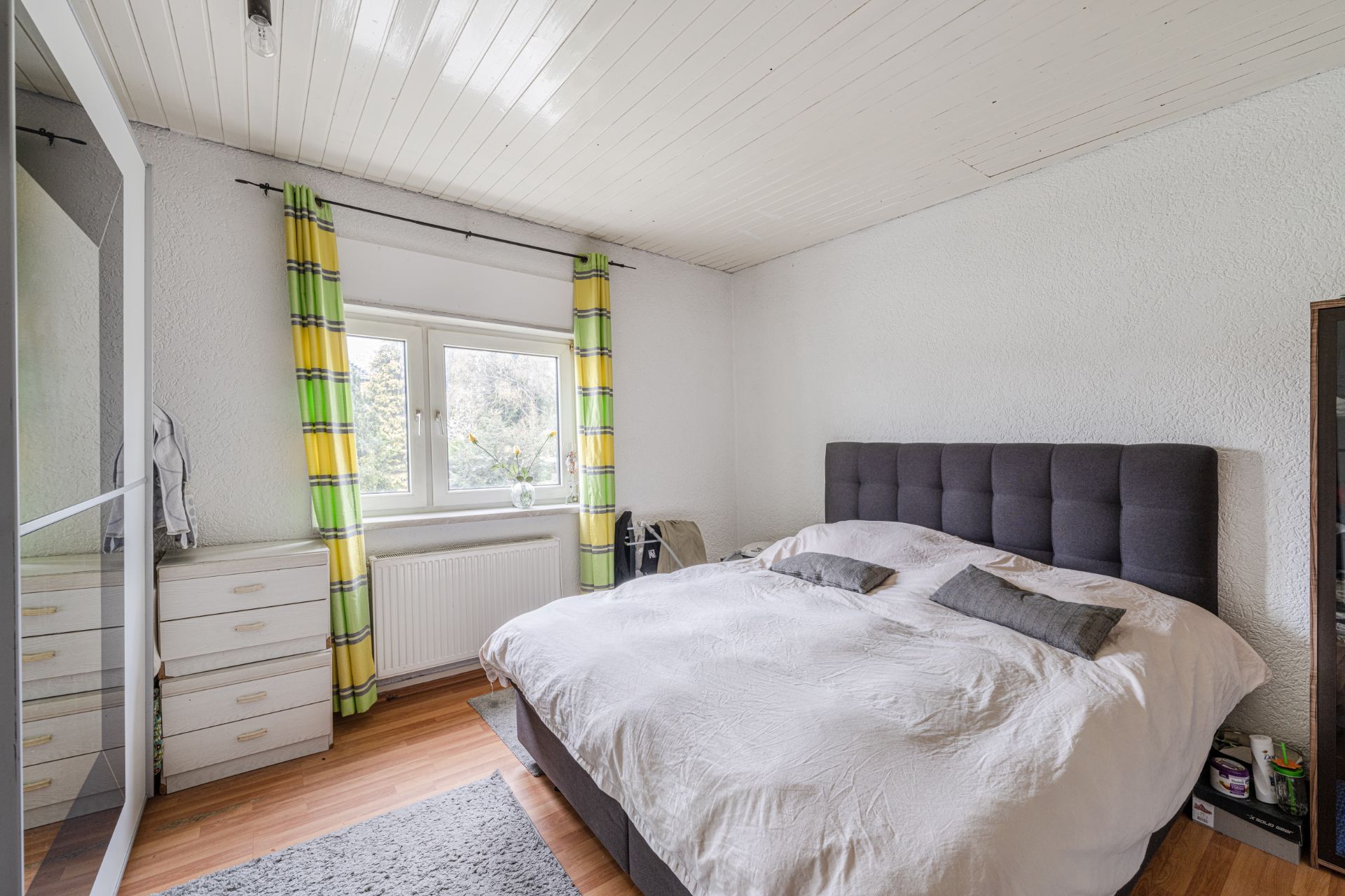 Helles Schlafzimmer mit großem Bett, gelb-grünen Vorhängen und Kommode am Fenster.