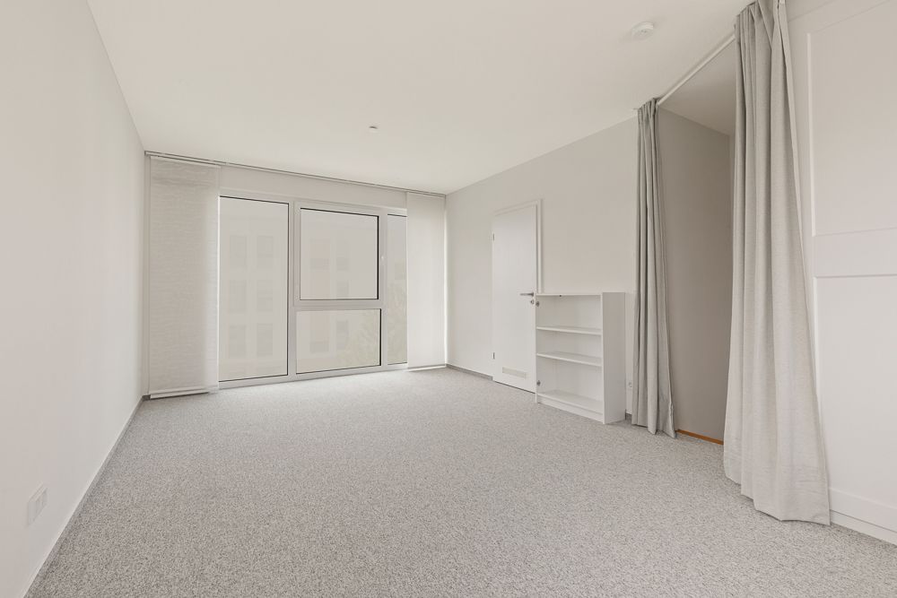 Helles, leeres Zimmer mit Teppichboden, großen Fenstern, Regal und Vorhang neben einer Tür.