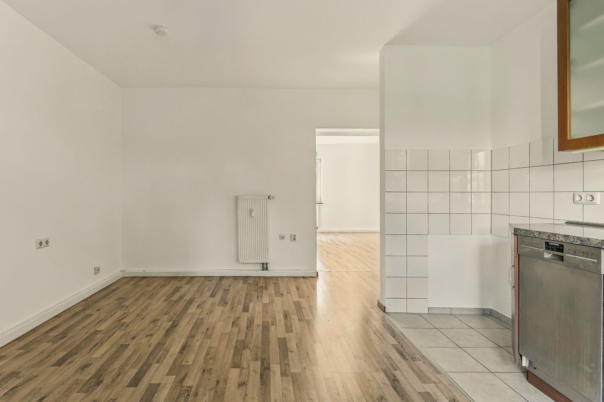 Leerer Raum mit Laminatboden, weißer Wand, Heizkörper und Küchenbereich mit Fliesen.