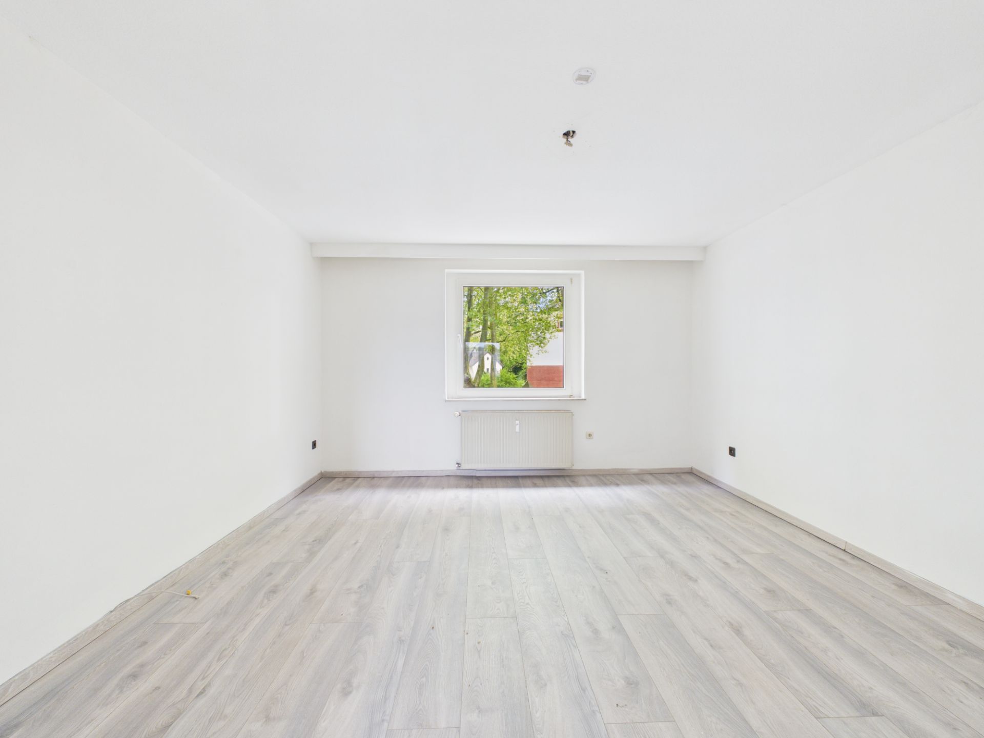 Leerer, heller Raum mit Fenster, weiß gestrichenen Wänden und hellem Laminatboden.