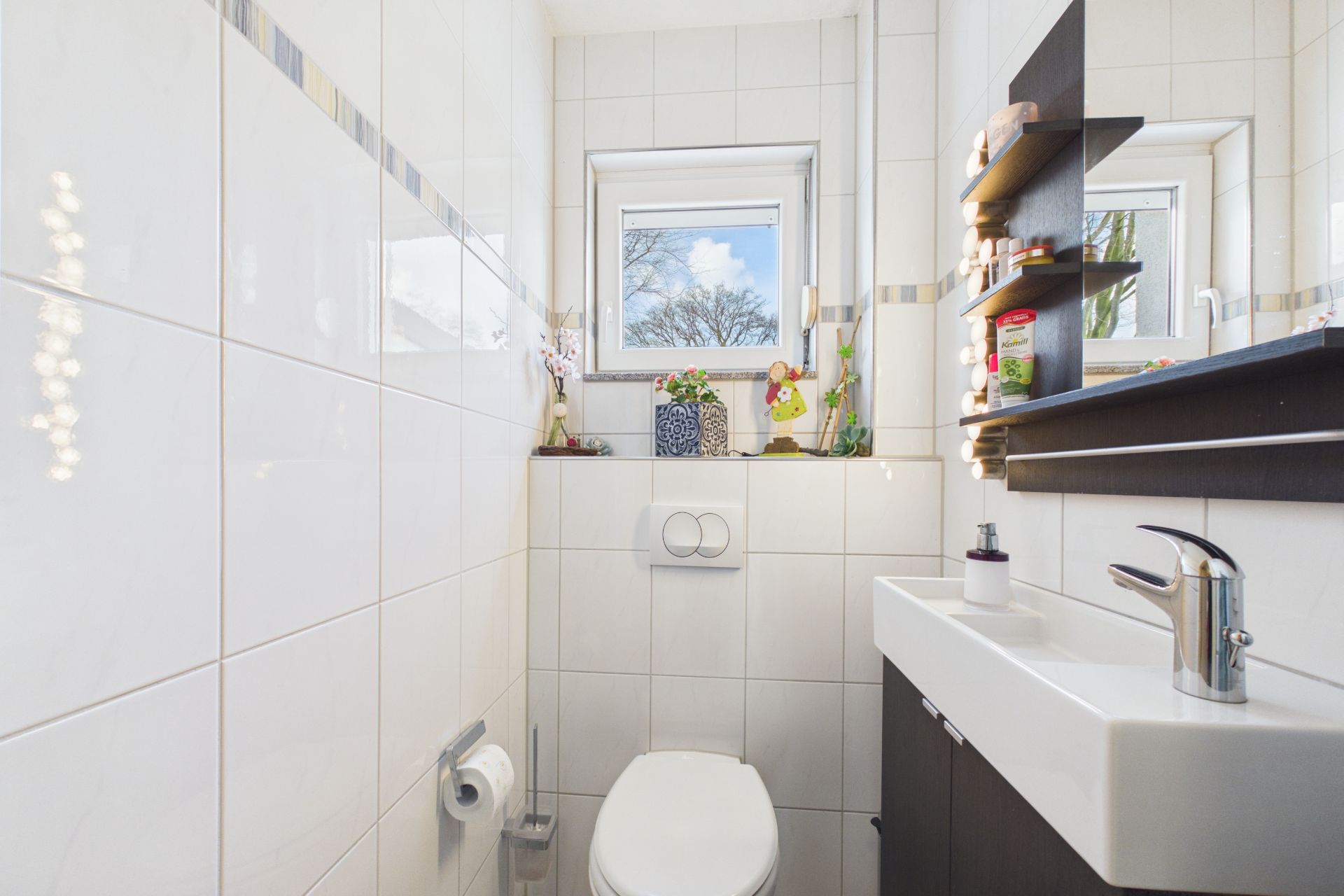Helles, kleines Bad mit Toilette, Waschbecken, Fenster und Regalen mit Dekoration.