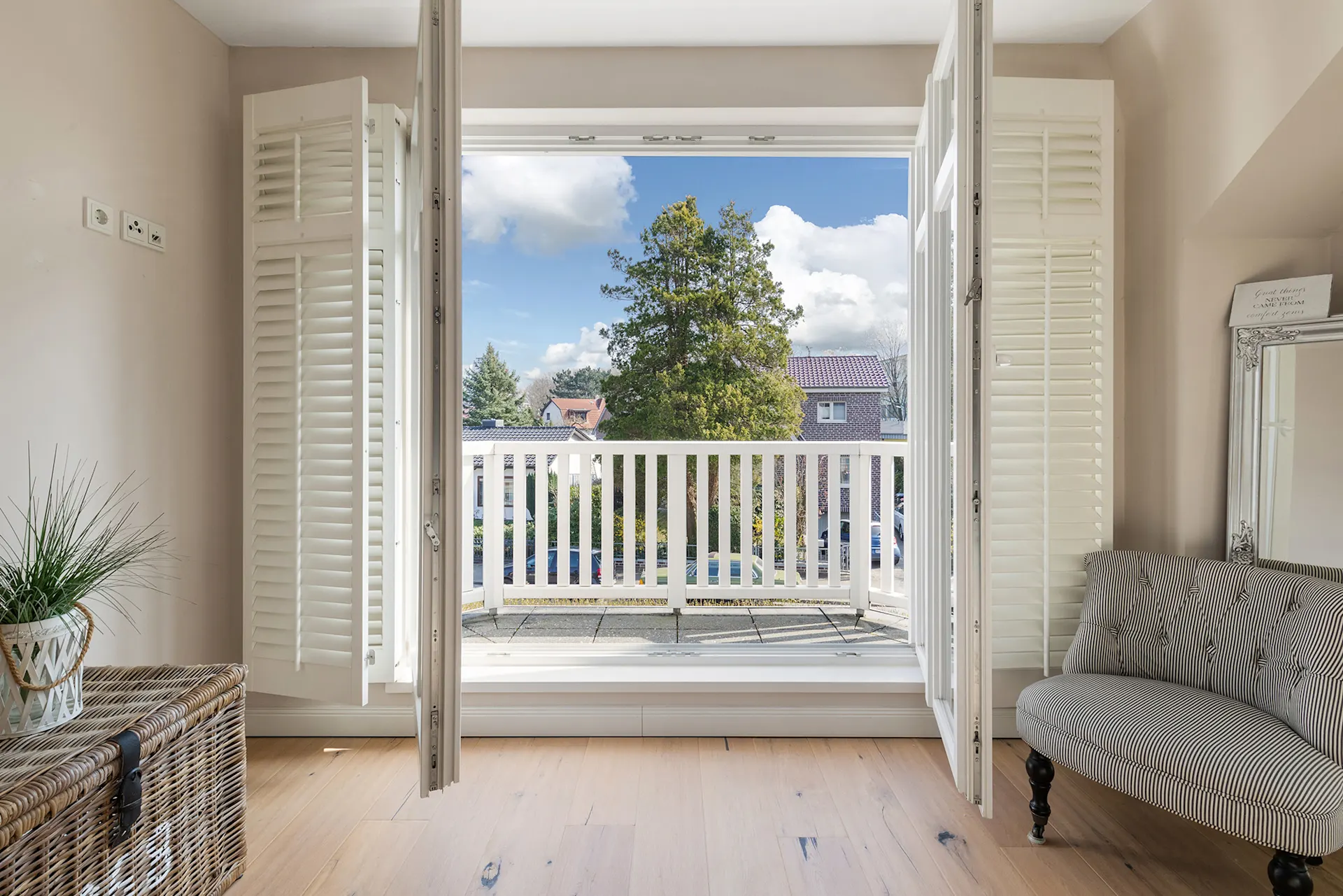 Helles Zimmer mit offenem Balkon, weißen Fensterläden, Sessel und Korb mit Pflanze.
