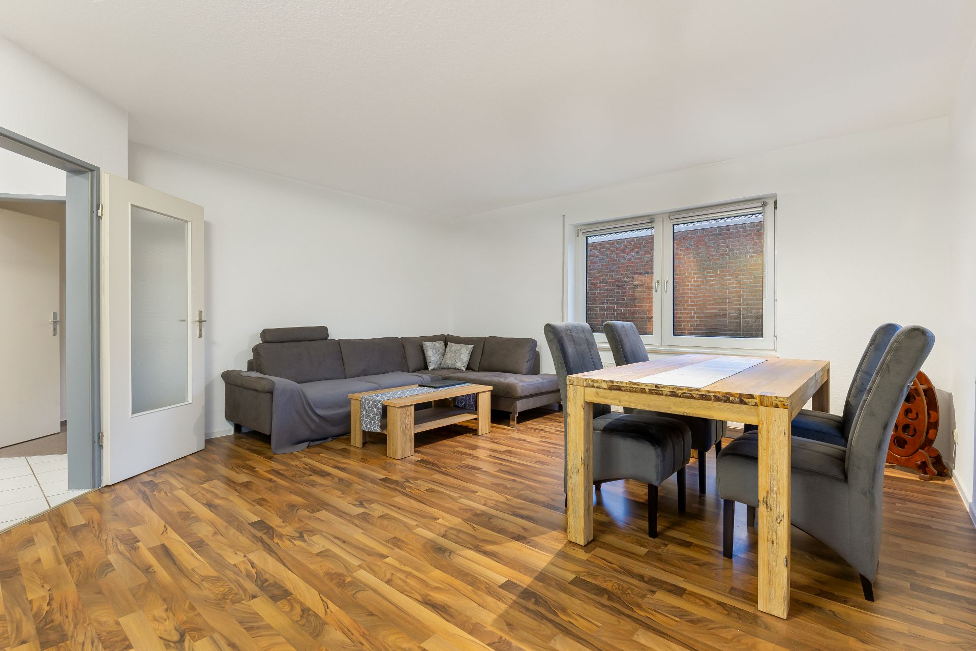 Wohnzimmer mit Holzboden, grauer Eckcouch, Esstisch und sechs Stühlen, Fenster an der Wand.