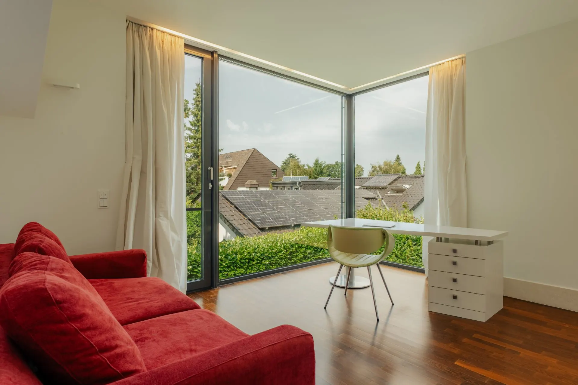 Wohnraum mit rotem Sofa, weißem Schreibtisch und großem Eckfenster mit Gartenblick.