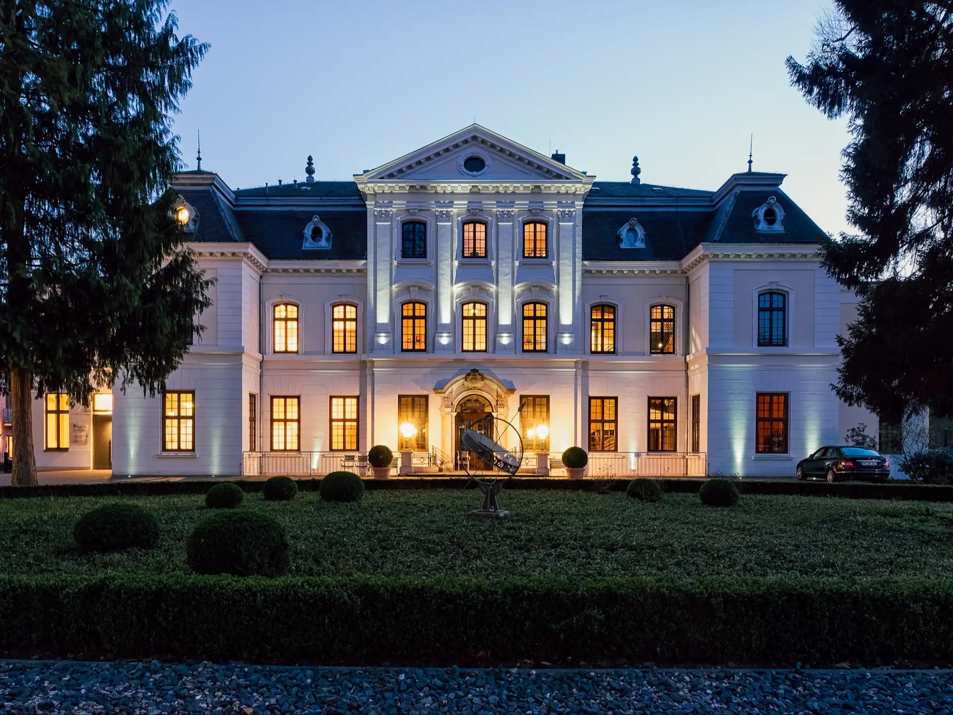 Großes weißes Wohngebäude mit vielen beleuchteten Fenstern und einem symmetrischen Garten im Vordergrund.