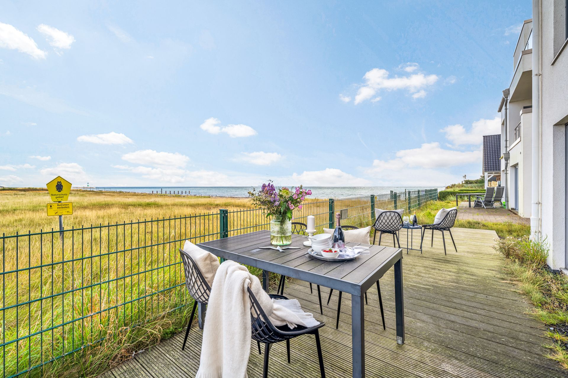 Terrasse mit Tisch und Stühlen, Blick auf eine Wiese und das Meer im Hintergrund.