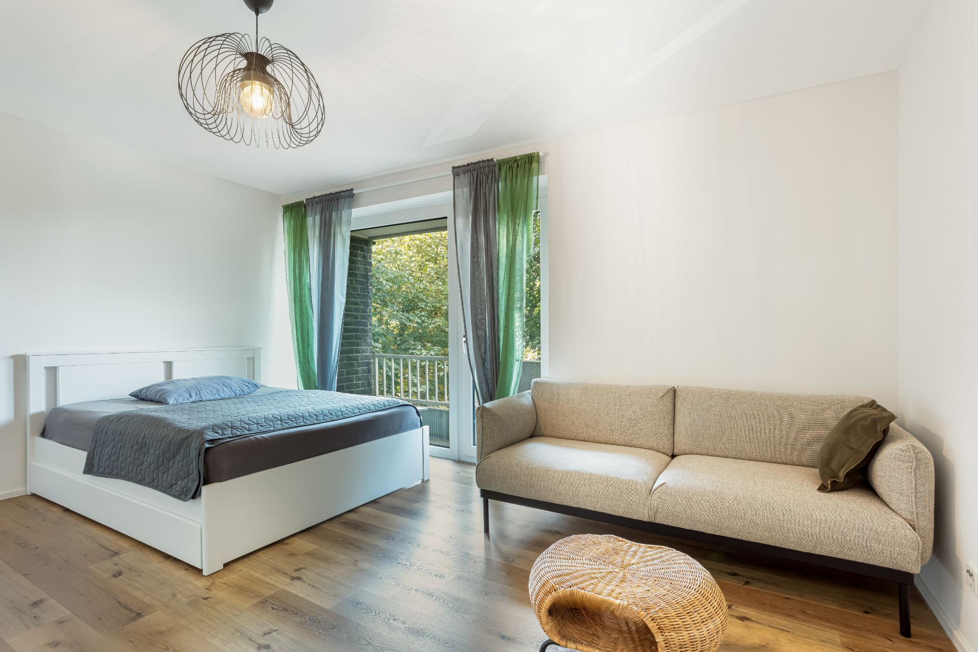 Helles Schlafzimmer mit Doppelbett, beiger Sofa und Zugang zu einem Balkon mit grünen Vorhängen.