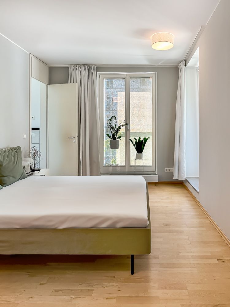 Helles Schlafzimmer mit Doppelbett, Holzboden, Fenstern, Vorhängen und zwei Pflanzen auf der Fensterbank.