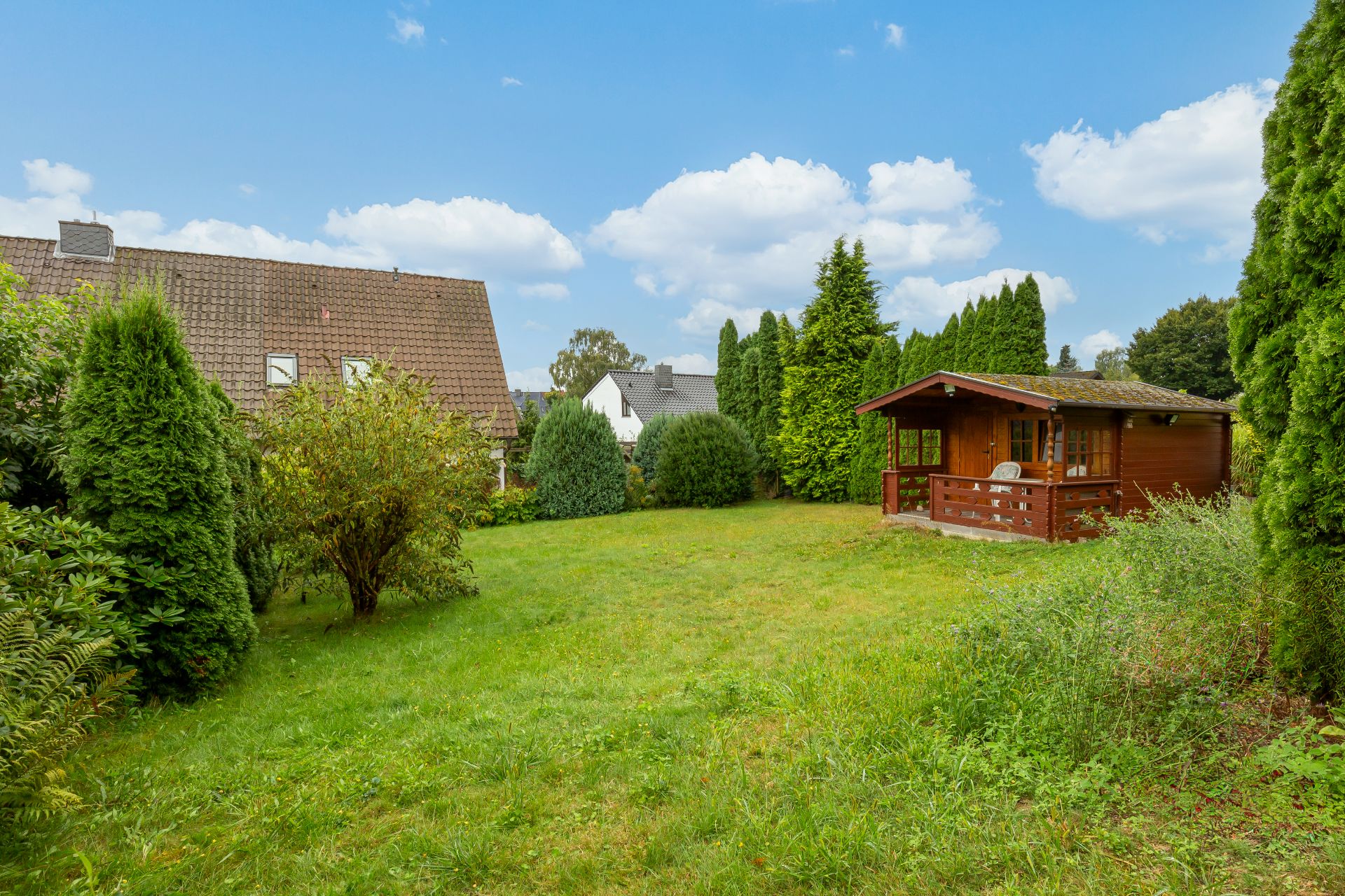 Grüne Wiese mit Büschen und Bäumen, rechts ein Holzhaus mit Veranda.